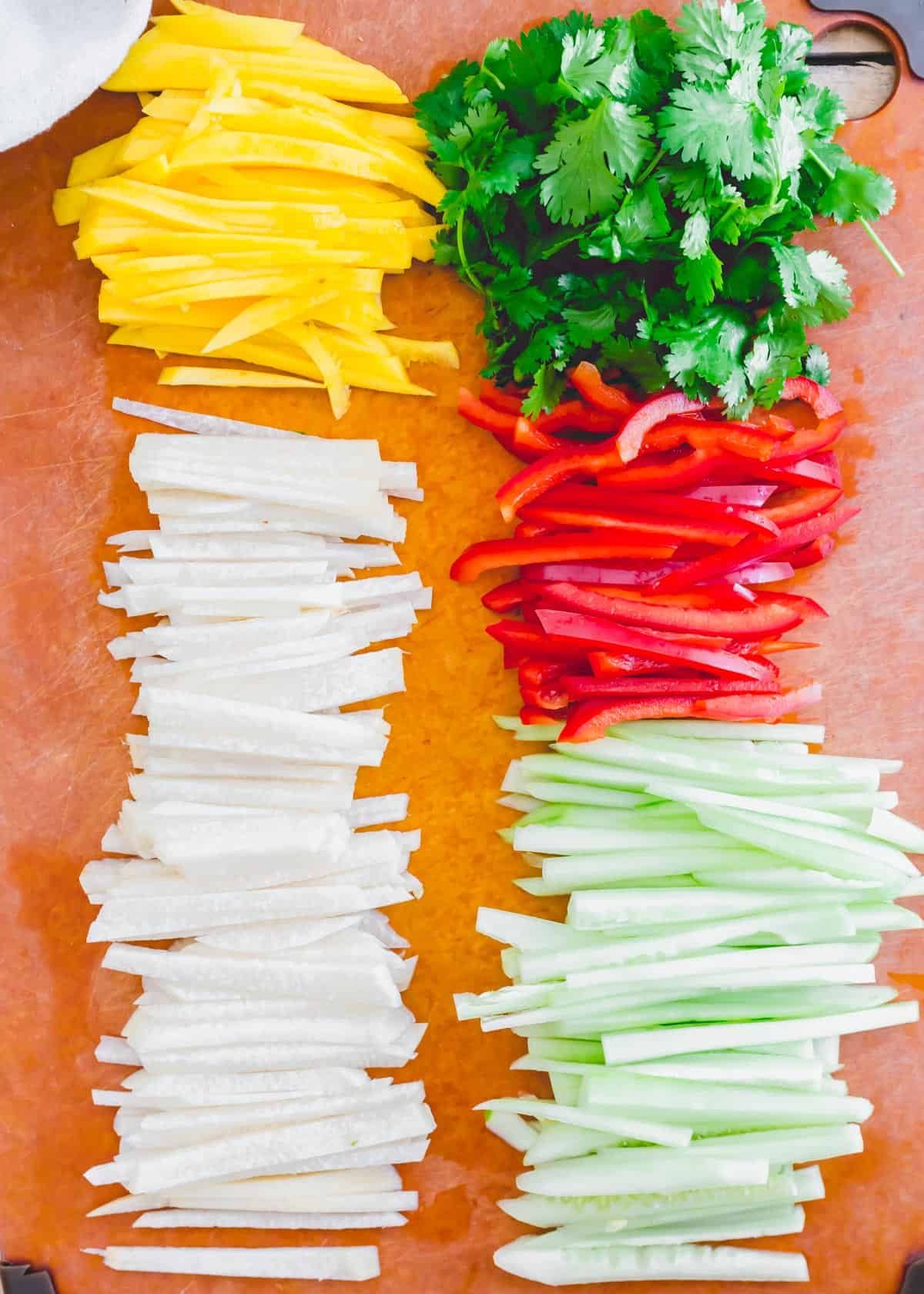 Julienned jicama, cucumber, red pepper and mango on a cutting board with fresh cilantro.