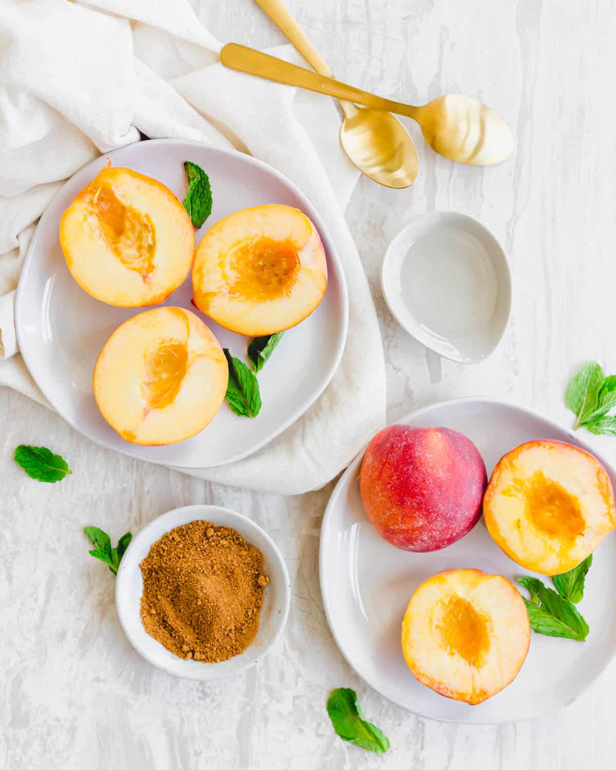 Halved yellow peaches on two white plates with fresh mint and cinnamon sugar on the side.