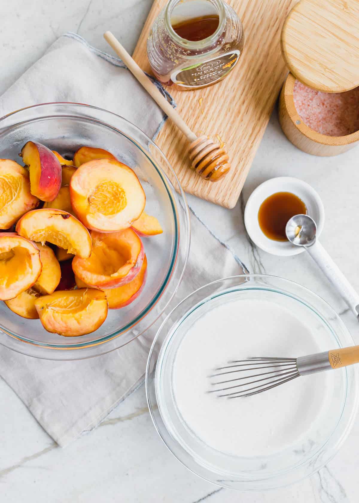 Fresh halved peaches in a glass bowl, coconut milk in another bowl and honey, vanilla and sea salt in the background.