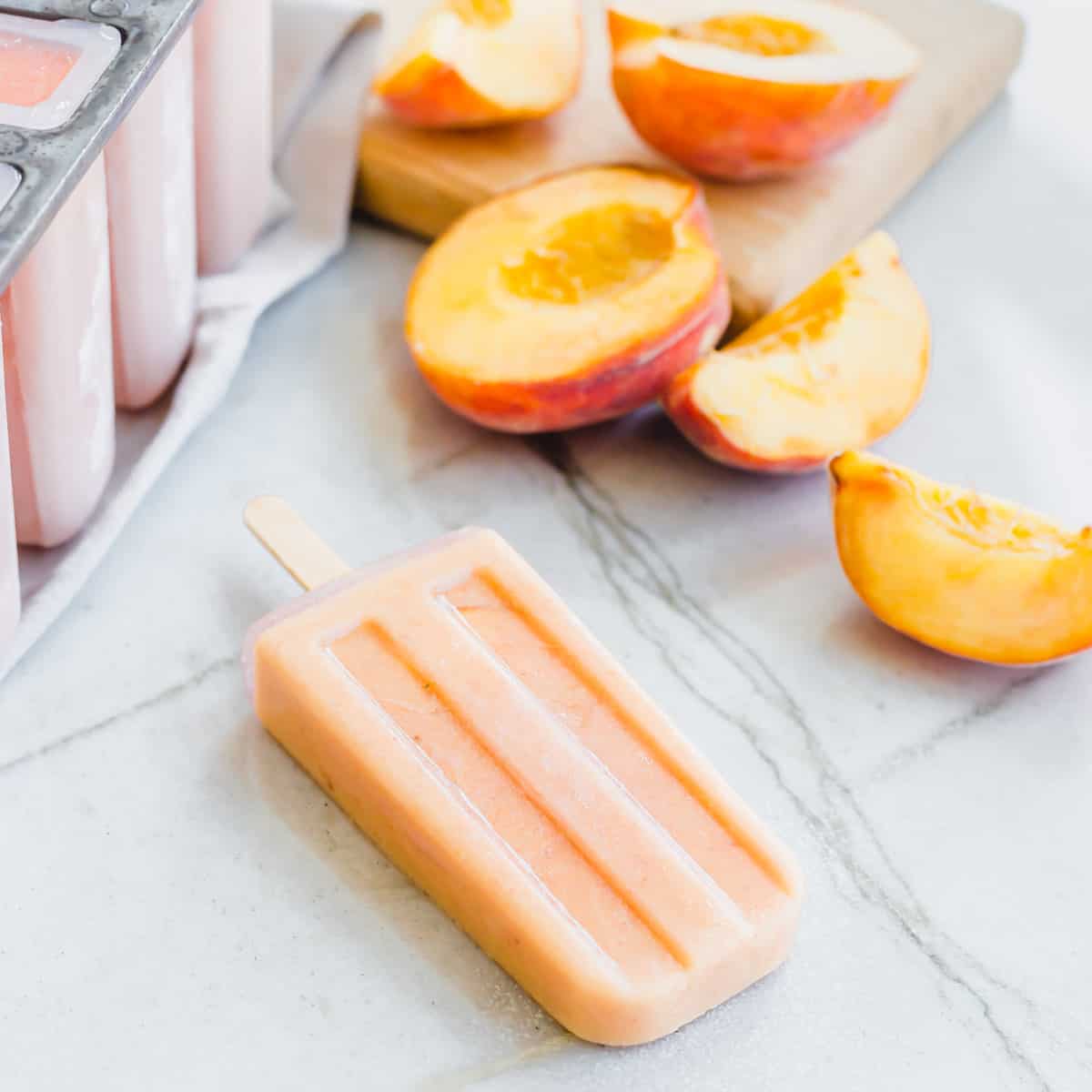 Dairy-free coconut peach ice pop on a marble surface with sliced fresh peaches in the background.
