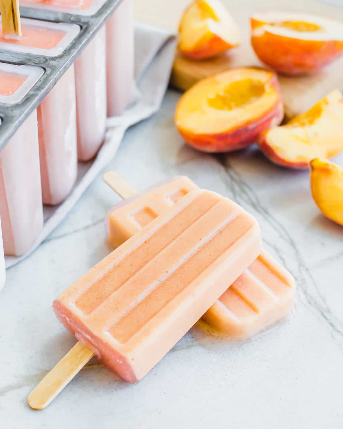 Creamy peach popsicles with peaches in the background on a marble surface.