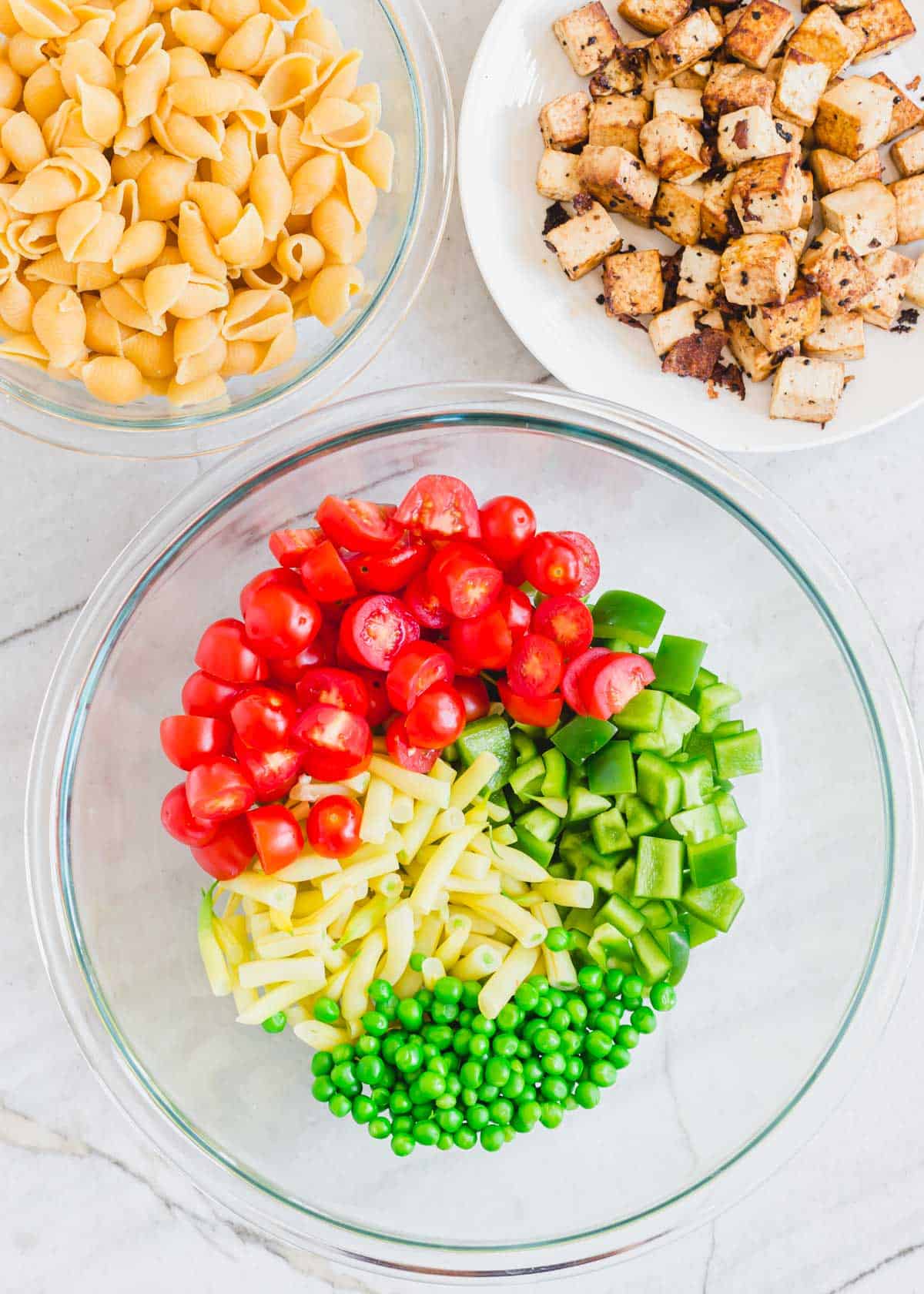 Ingredients to make a tofu pasta salad recipe including cherry tomatoes, green peppers, peas and yellow wax beans in a glass bowl with cooked tofu on a plate and cooked shell pasta in another bowl.