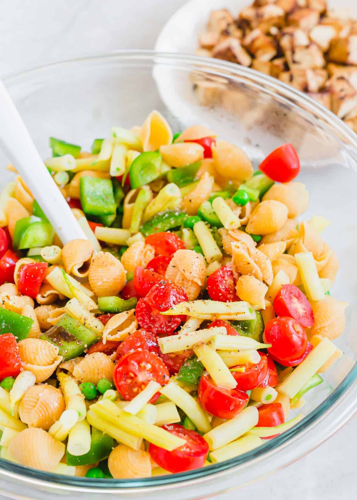 Pasta, tomatoes, yellow wax beans, peas and peppers in a glass bowl with salt, pepper and a spatula.