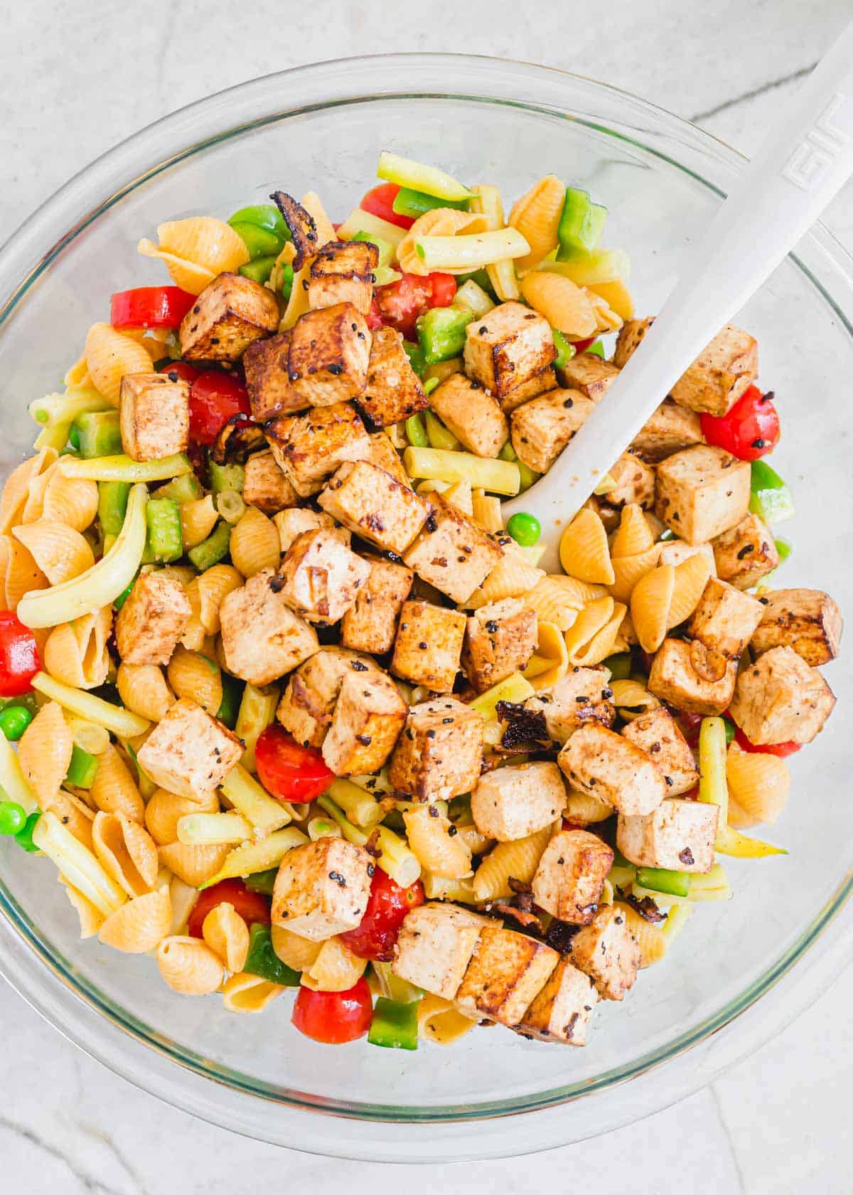 Skillet cooked tofu on top of pasta salad in a glass bowl with a white spatula.