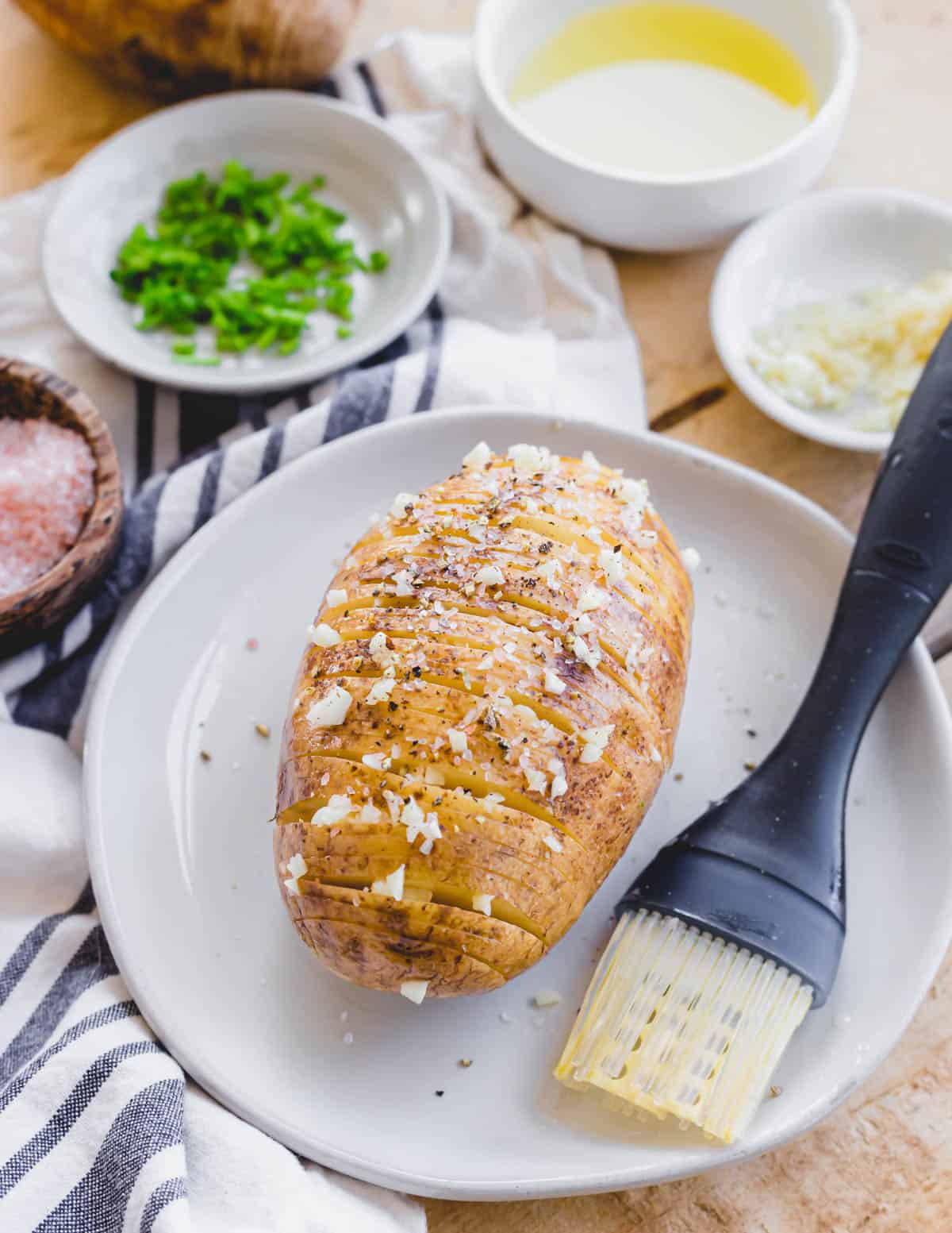 A russet potato thinly sliced in hasselback fashion seasoned with ghee, salt, pepper and minced garlic on a plate with a silicone brush.