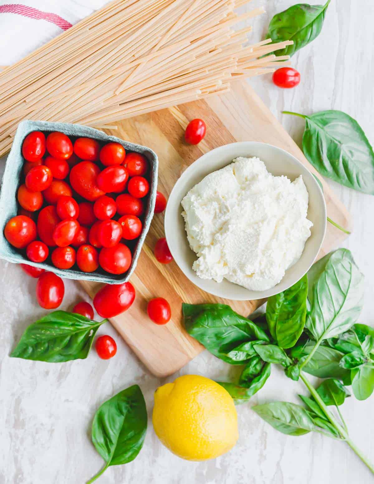 Ingredients to make ricotta lemon pasta: cherry tomatoes, fresh ricotta, basil and linguine on a cutting board.