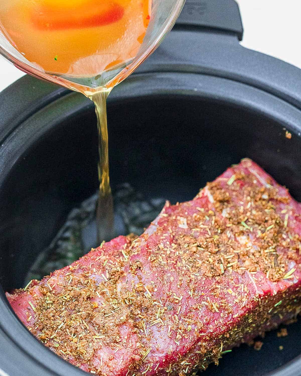 Seasoned tri tip roast in a slow cooker with broth being poured in.