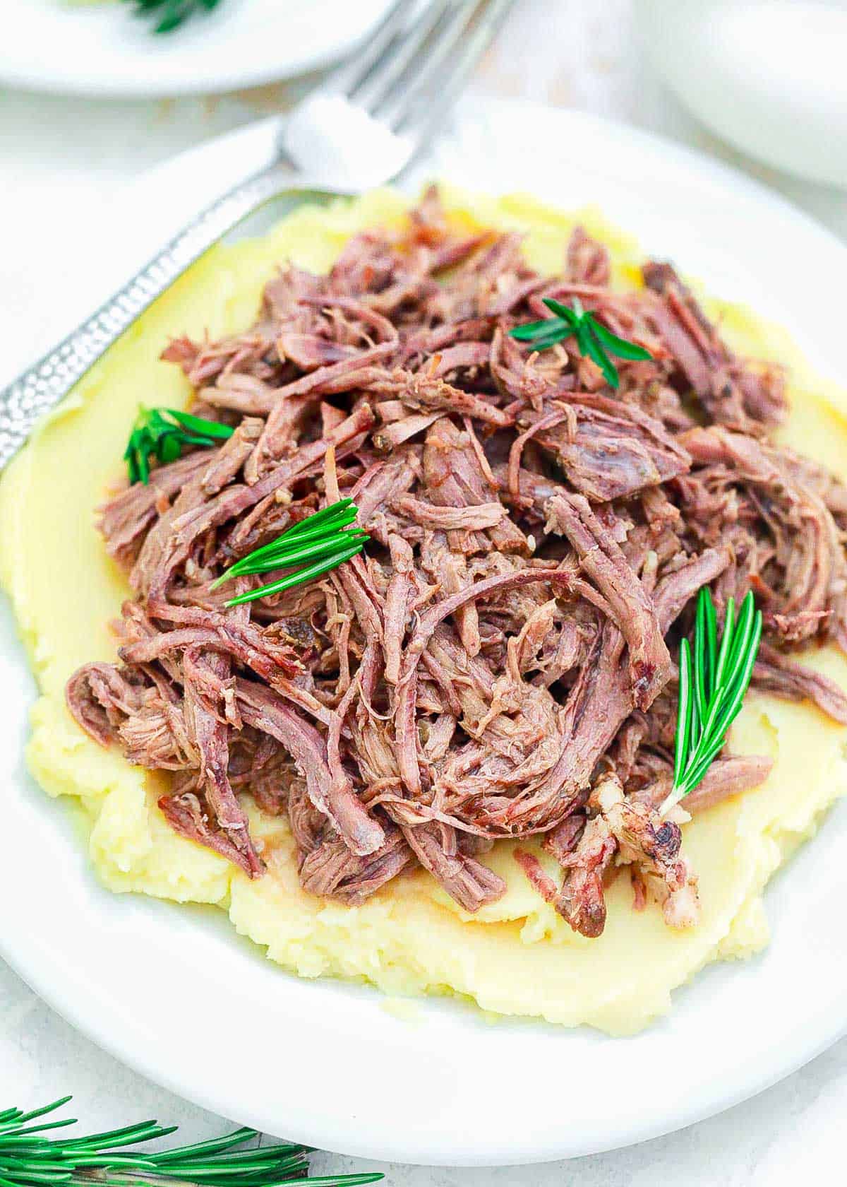 Shredded slow cooker tri tip recipe served over mashed potatoes garnished with fresh rosemary.