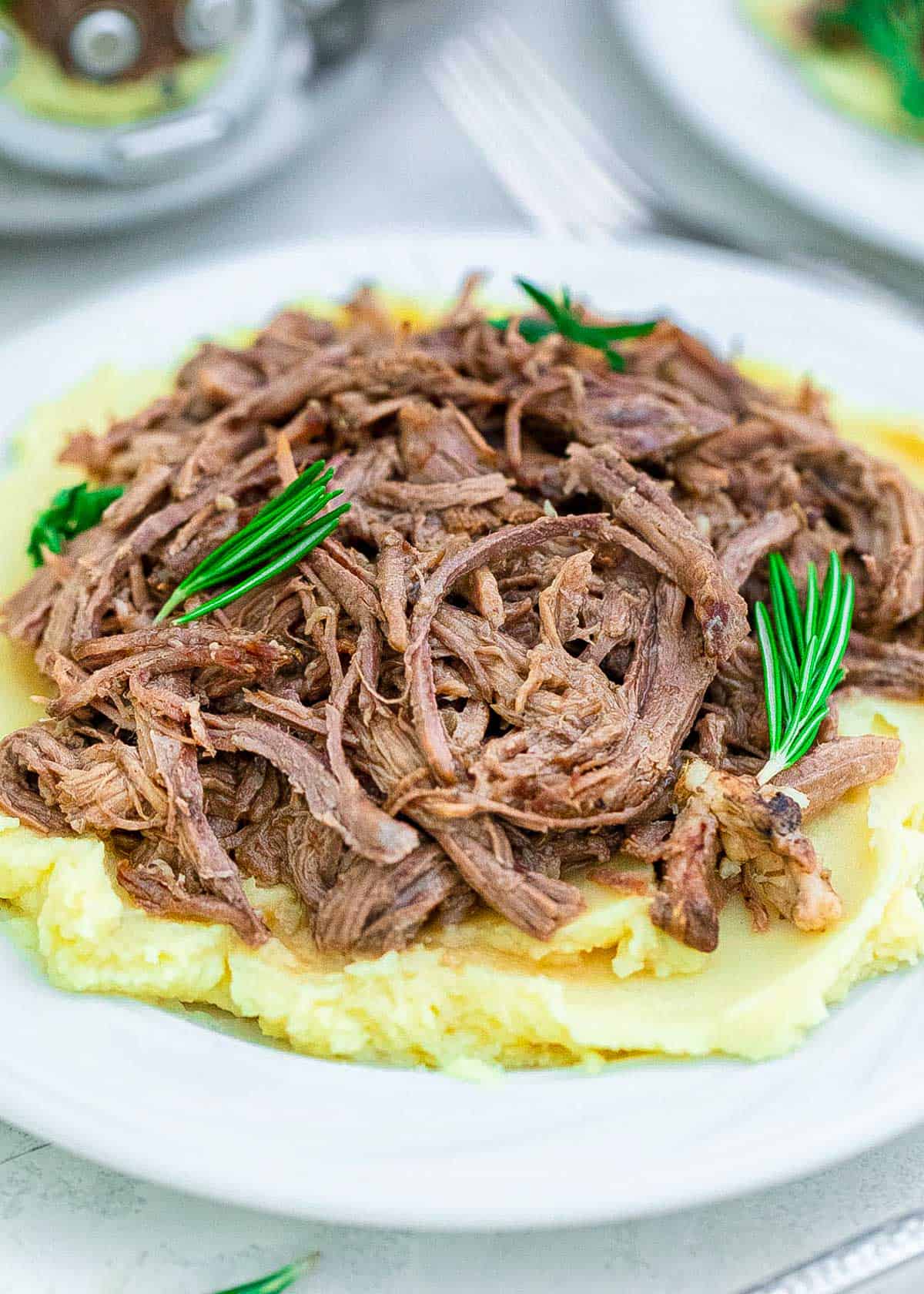 Crockpot tri tip roast shredded and piled on top of mashed potatoes on a white plate with fresh rosemary garnish.