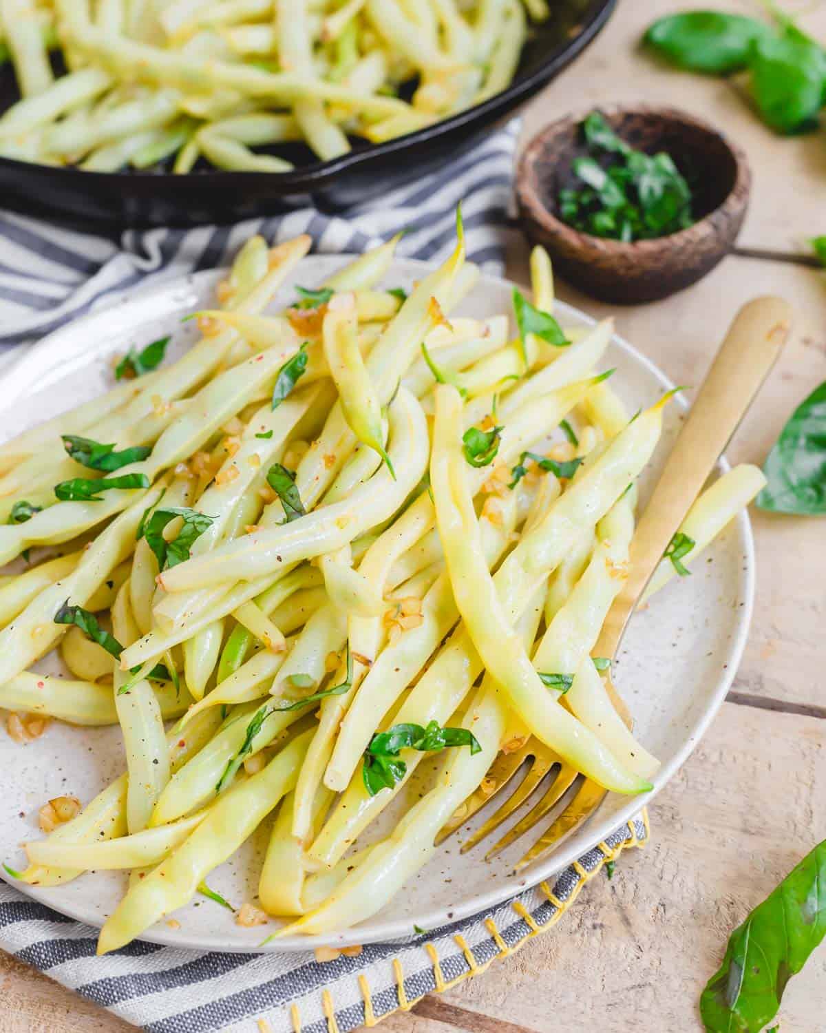 Cooked wax beans on a plate with sautéed garlic and fresh chopped basil with a fork on the side.