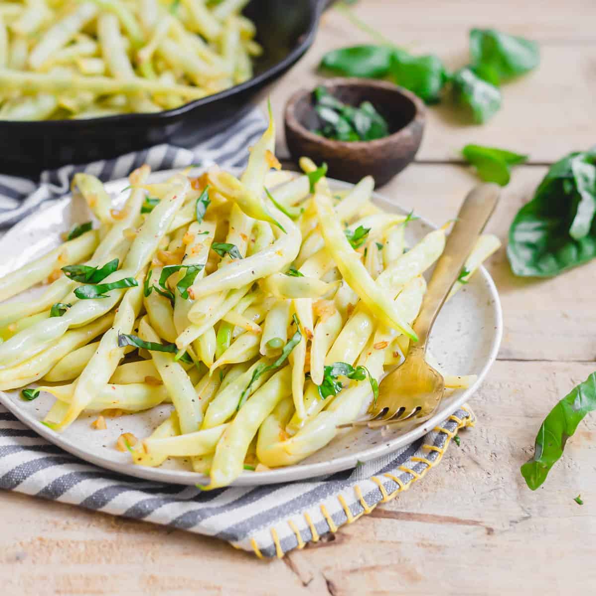 Cooked yellow beans with garlic and basil on a plate with a gold fork and striped napkin.