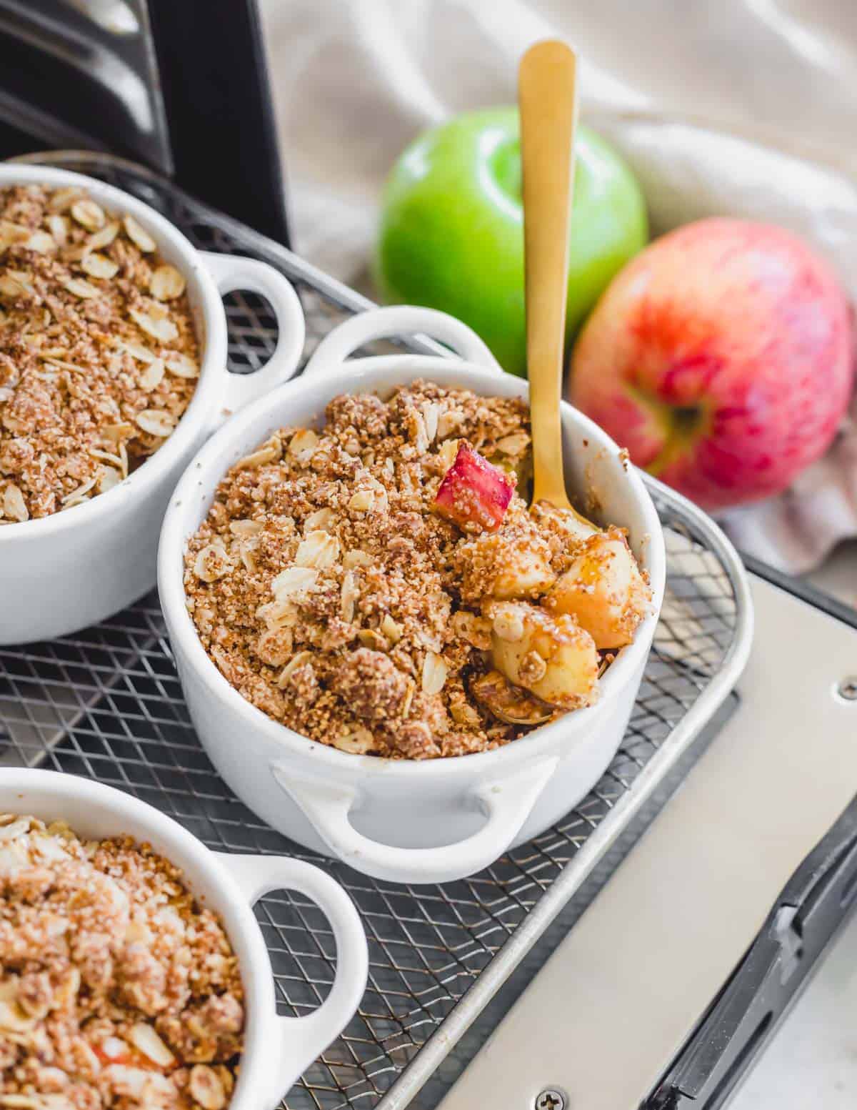 Healthier apple crisps in ramekins on an air fryer tray with a spoon.