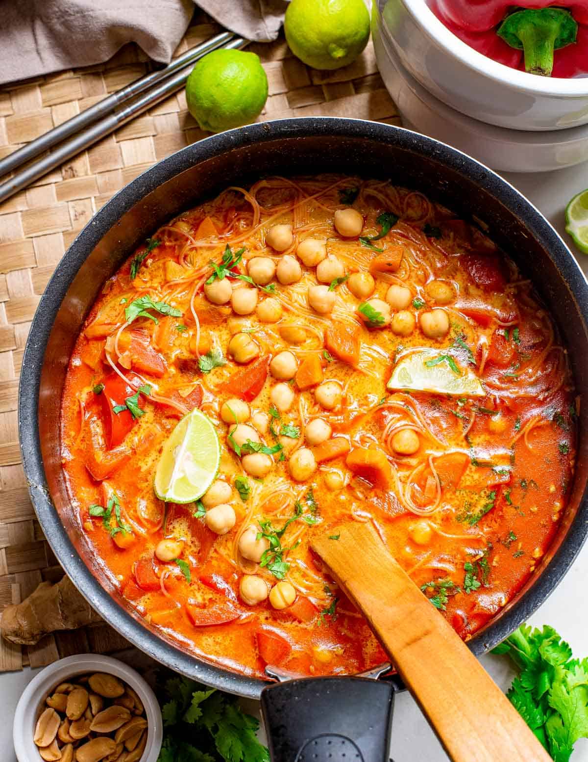 Red curry noodle soup with lime wedges and cilantro in a pot with a wooden serving spoon.