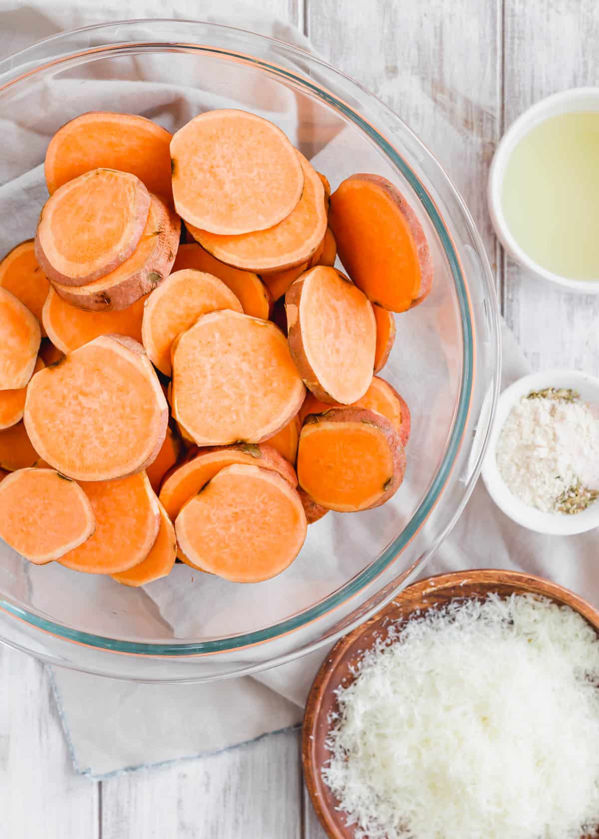 Sweet potato rounds in a large glass bowl with oil, herbs and parmesan cheese in dishes on the side.