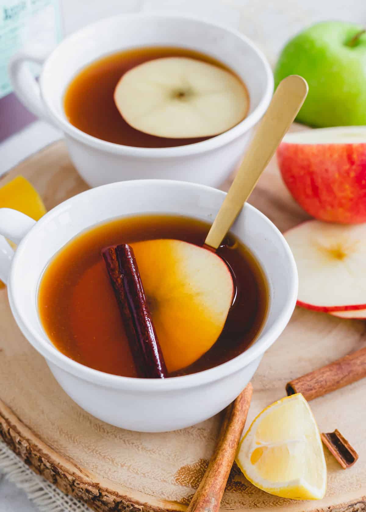 Apple cider tea with an apple slice and cinnamon stick in a white mug with a gold spoon.