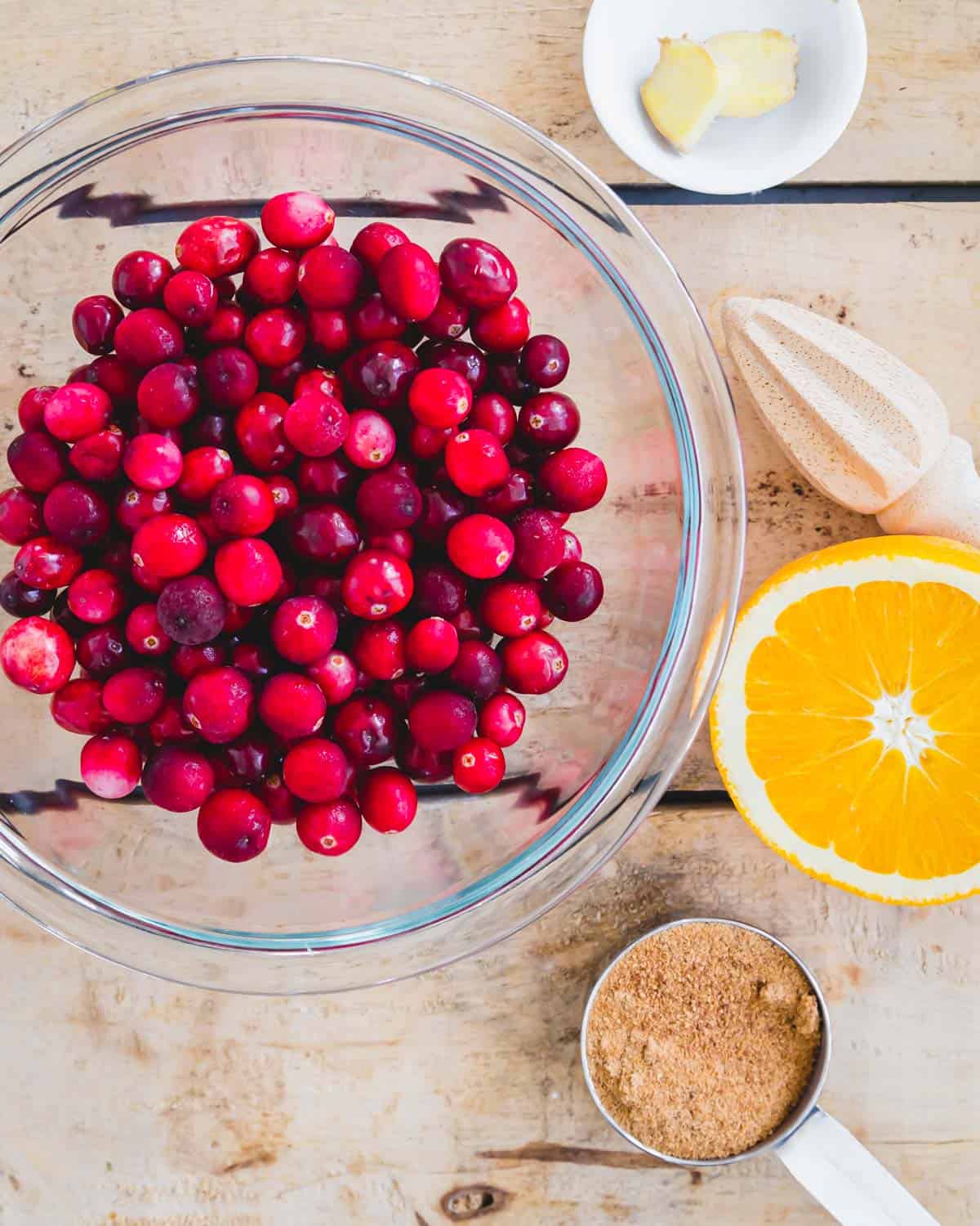 Fresh cranberries in a glass bowl with half an orange, fresh ginger slices and coconut sugar on a wooden surface.