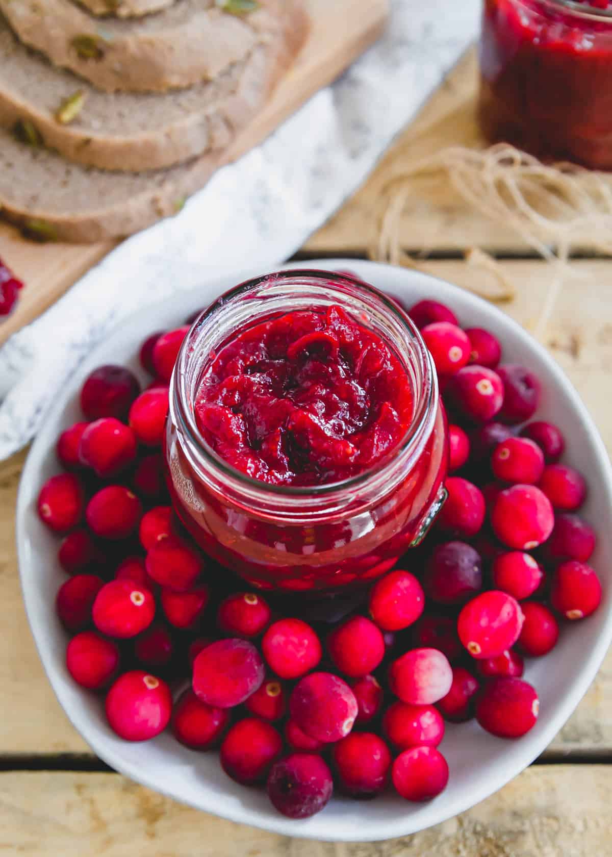 Cranberry jam in a glass jelly jar surrounded by fresh cranberries on a plate.