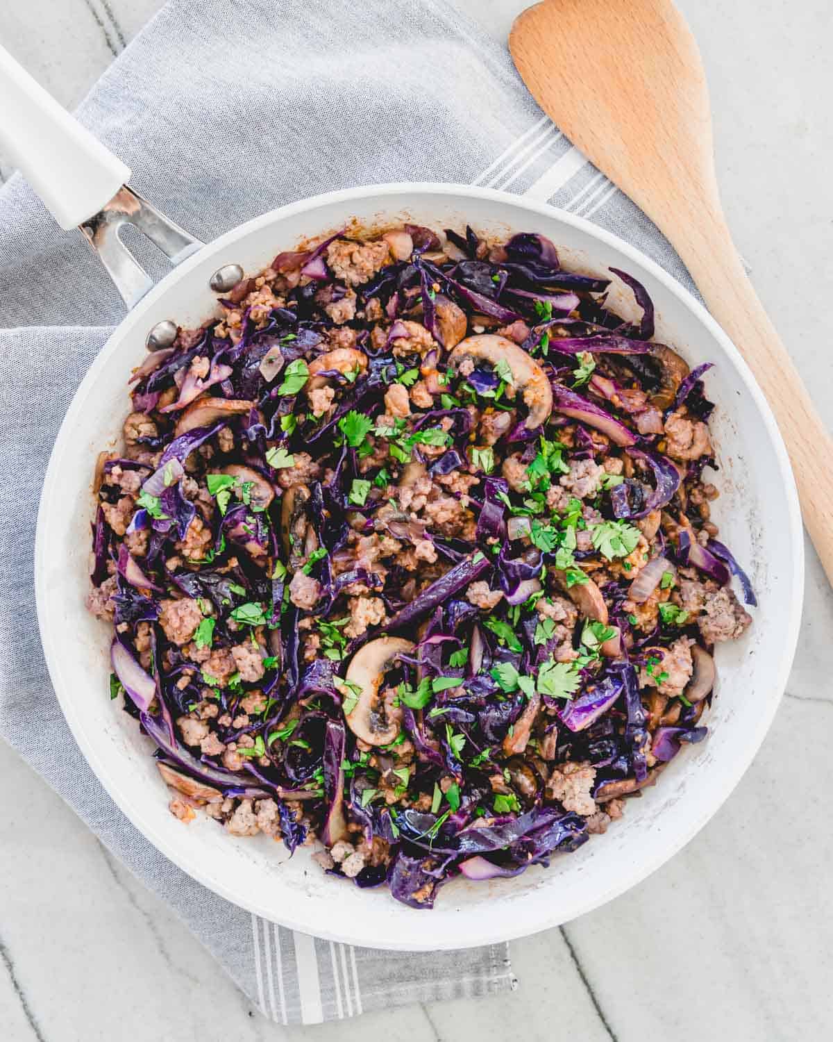 Pork and cabbage stir fry in a white skillet on a gray cloth napkin with a wooden spatula on the side.