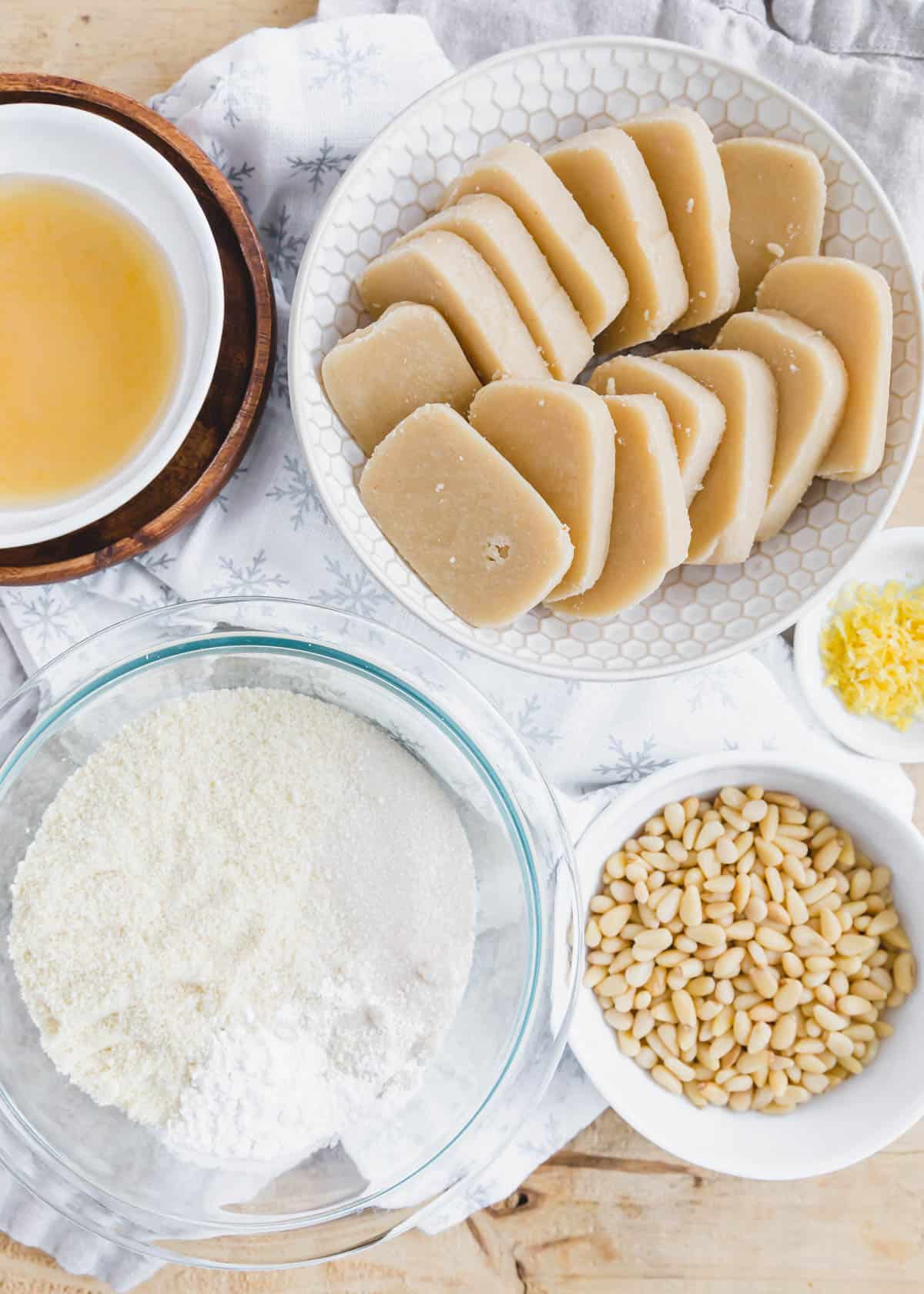 Ingredients to make pignoli cookies without egg whites in bowls including almond paste, lemon zest, pine nuts and a flour mixture.