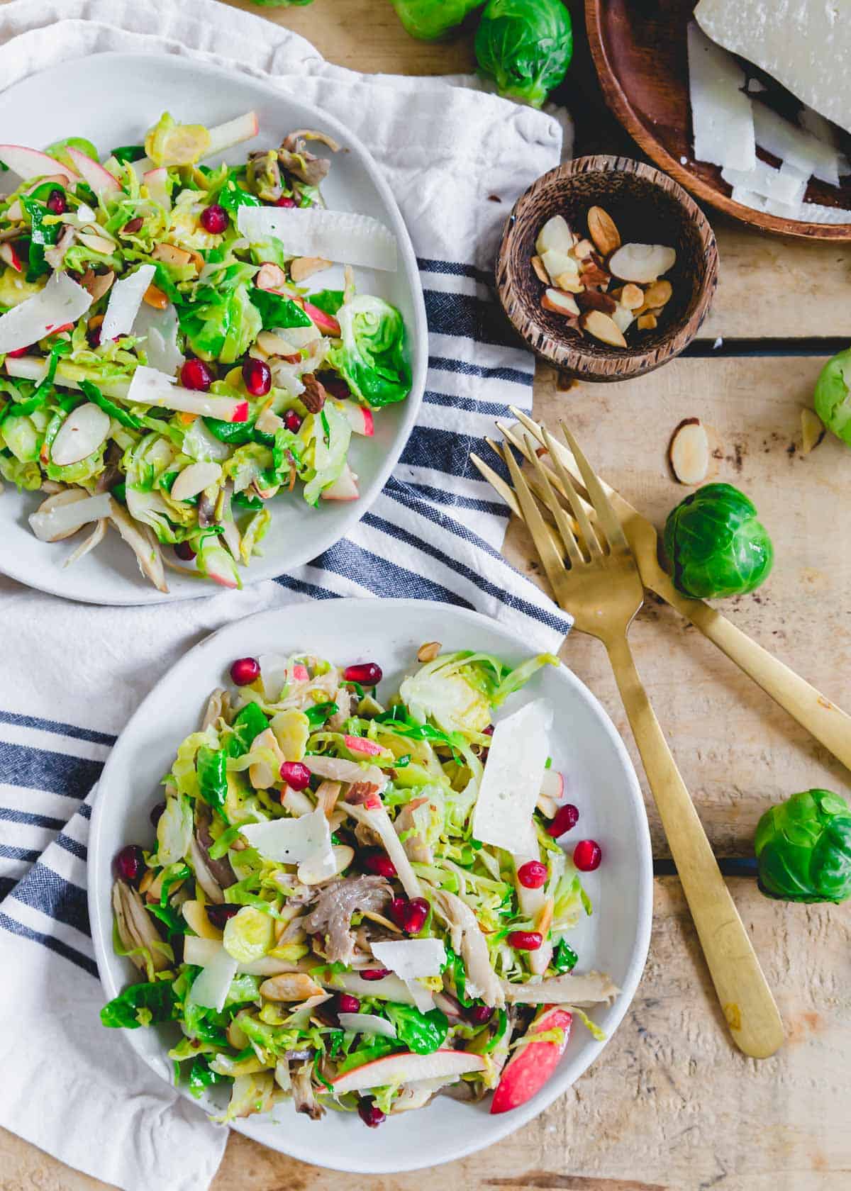Shredded Brussels sprouts salad with chicken, apples, pomegranates, sliced almonds and pecorino cheese on a white plate with gold fork on the side.