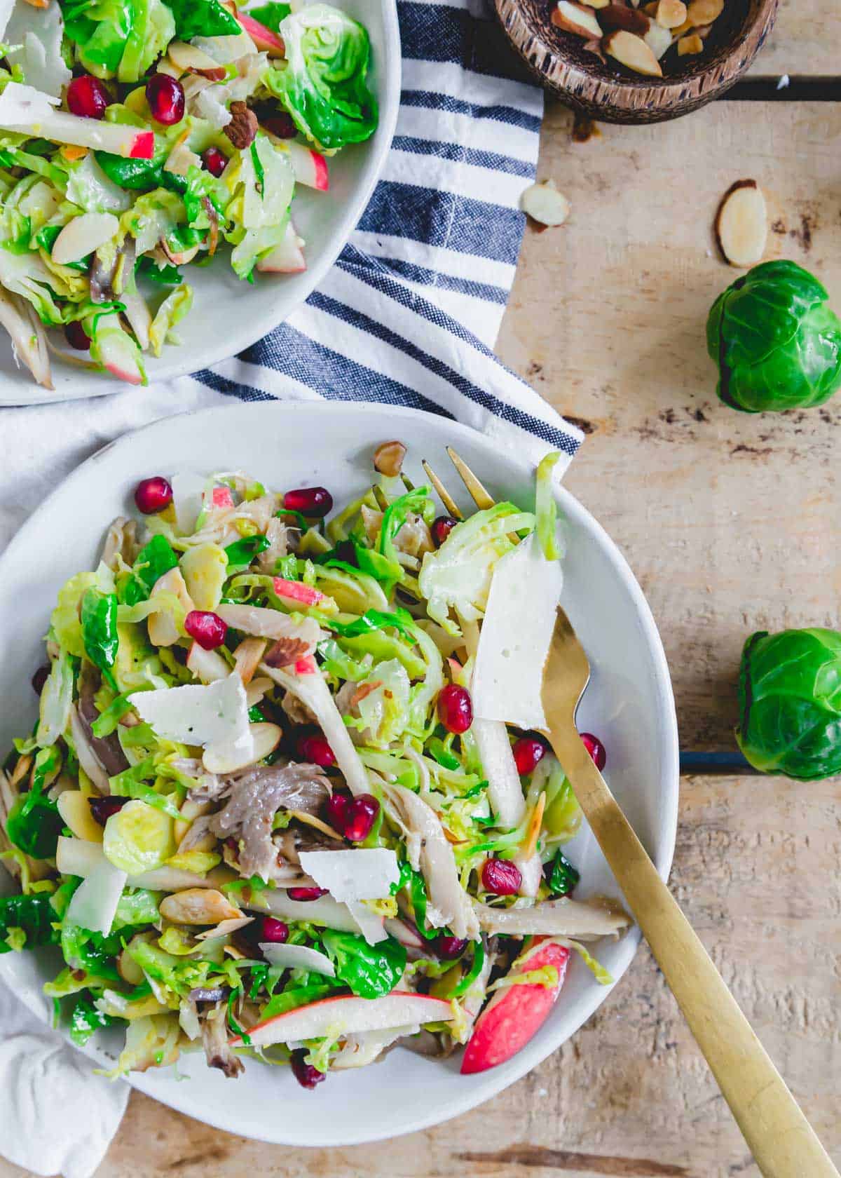 Shredded chicken and Brussels sprouts salad recipe with apples and pomegranates tossed in a honey mustard dressing on two white plates.