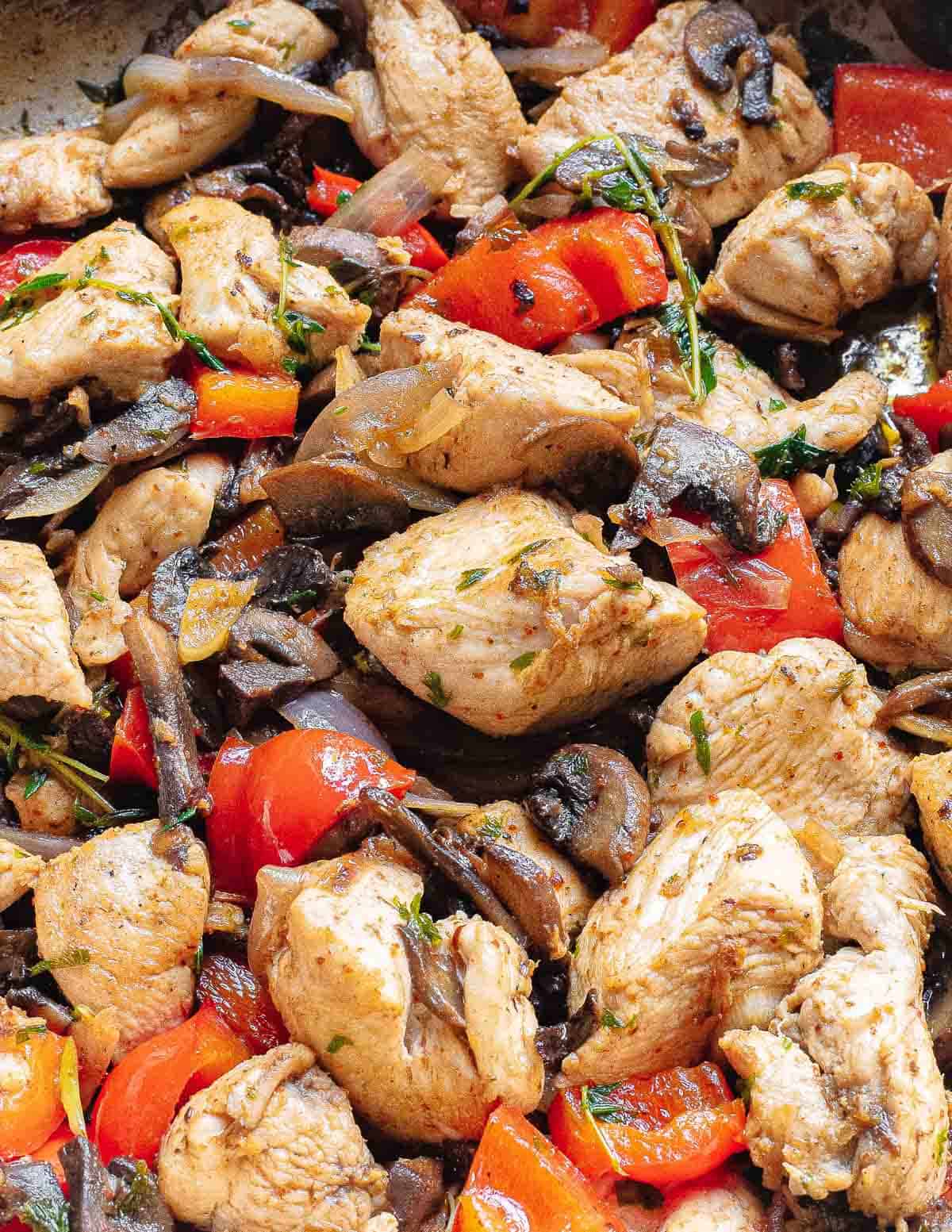 Browned chicken breast bites in a skillet with red bell peppers, mushrooms, onions and fresh herbs.