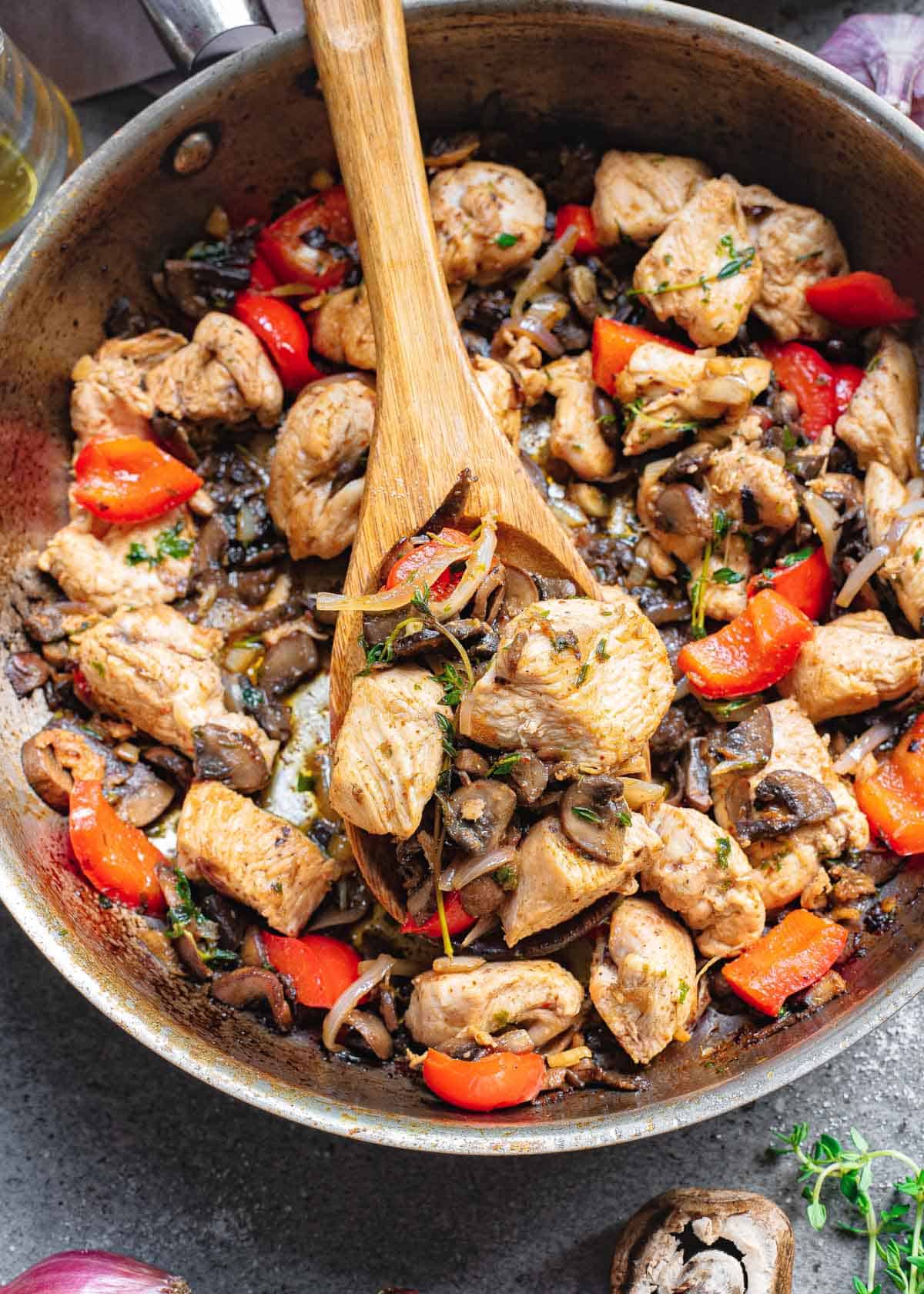 One-pan, 20 minute chicken bite dinner with garlic, mushrooms, red peppers and onions in a skillet with a wooden spoon.