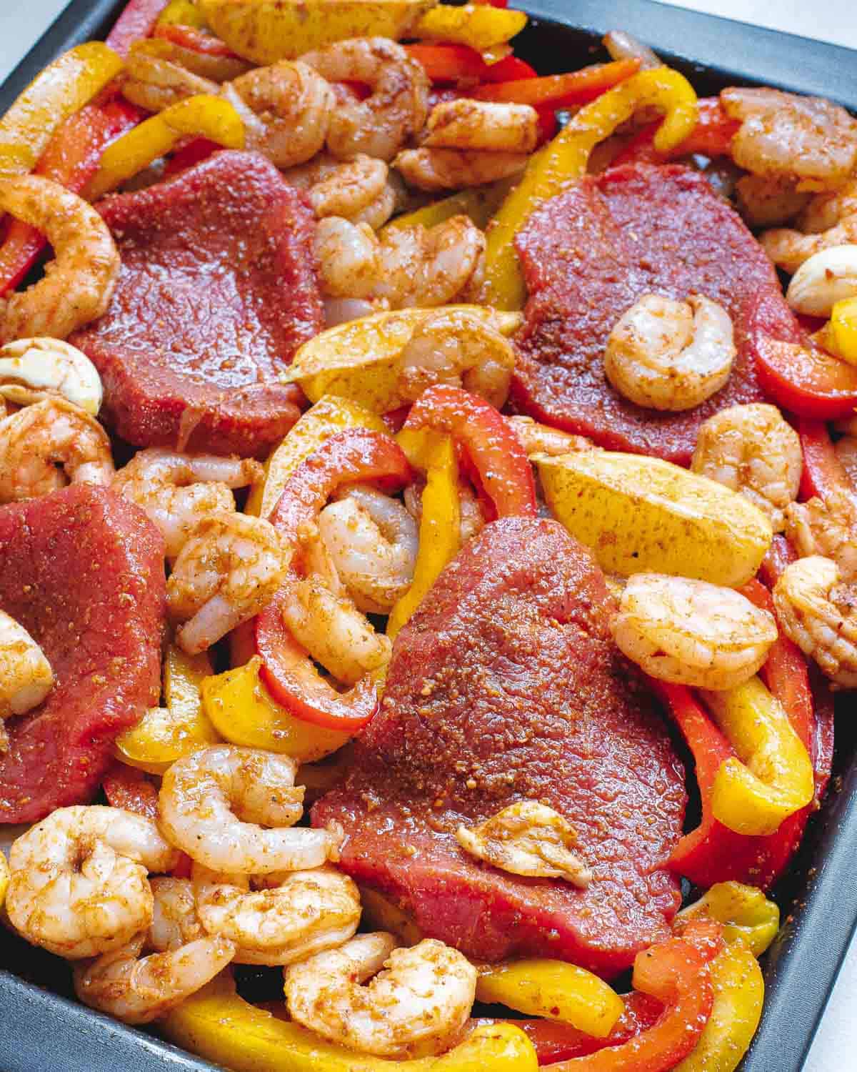 Sirloin steak, shrimp, peppers and onions tossed in fajita seasoning on a baking sheet.
