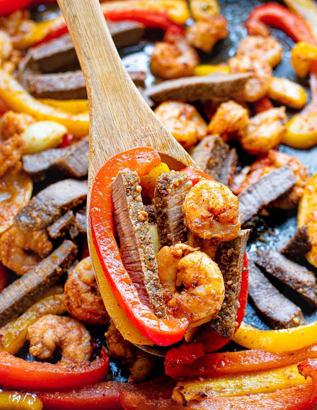 Shrimp and steak fajitas on a sheet pan with a wooden spoon.