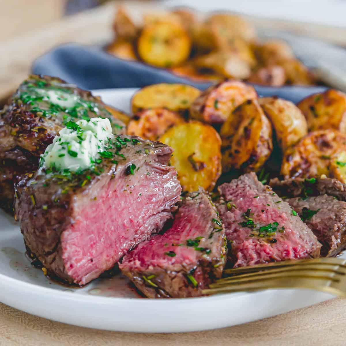 Medium rare filet mignon with a gold fork and baby potatoes on a plate with fresh herbs.