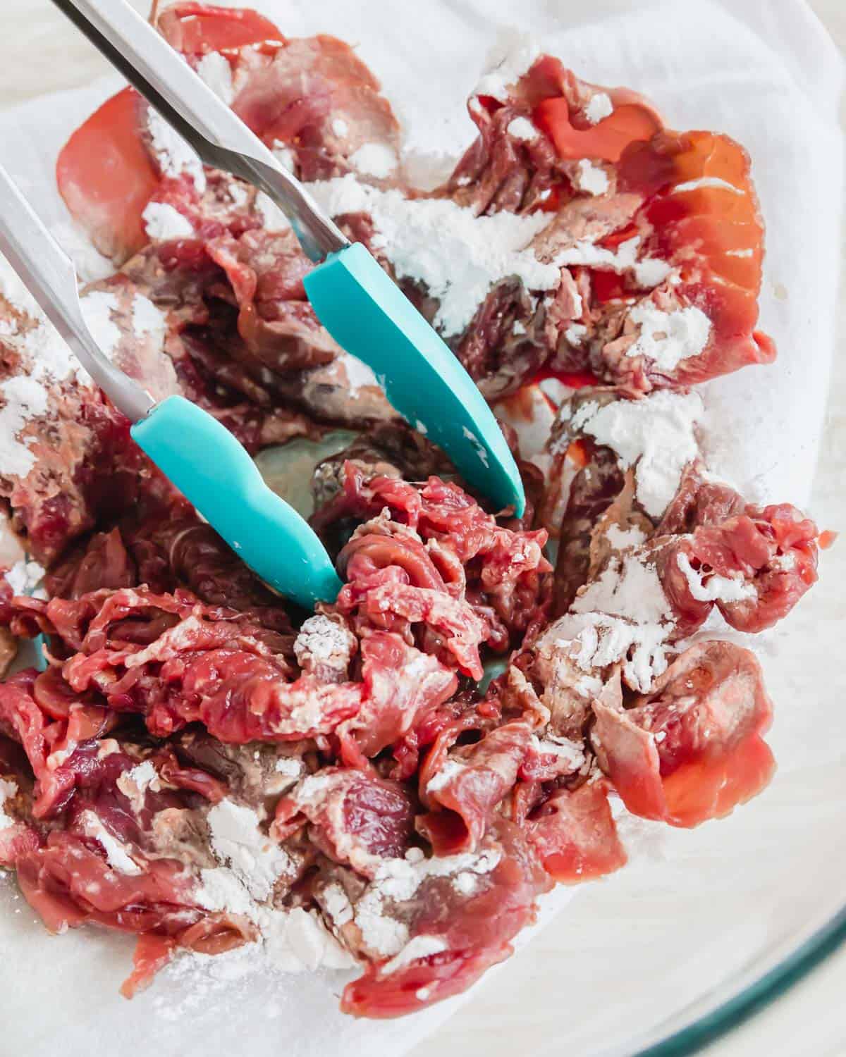 Shaved beef steak tossed with arrowroot powder in a bowl with tongs.