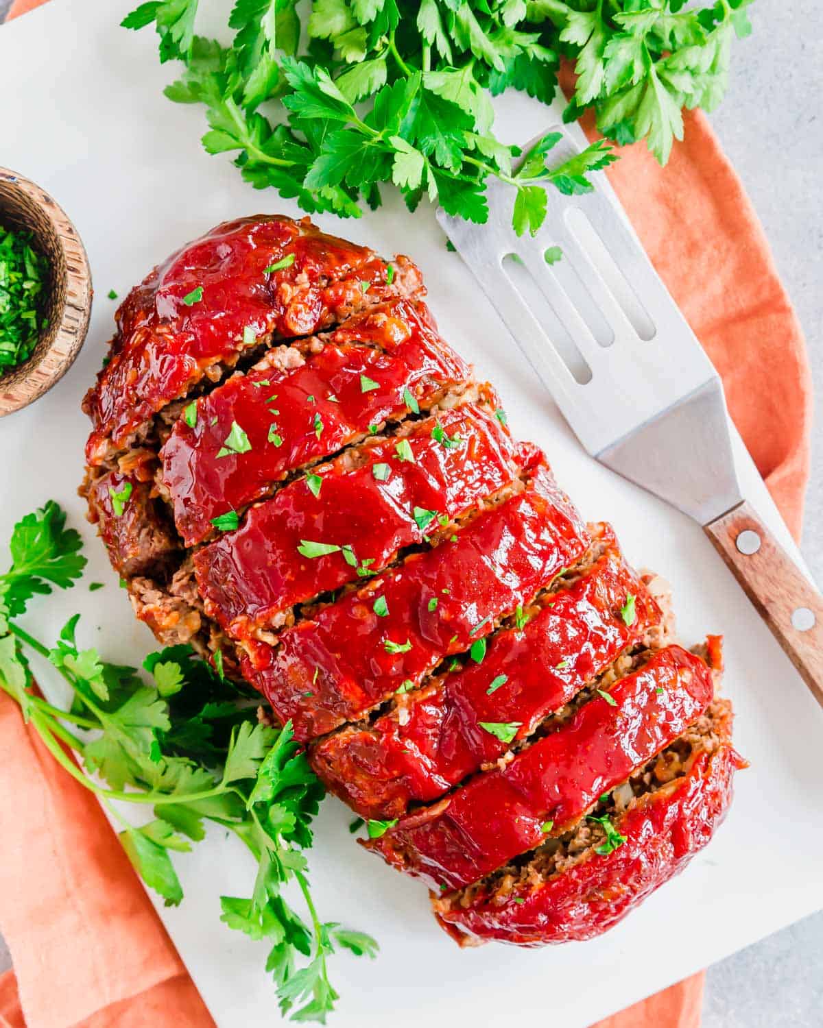 Cooked meatloaf sliced on a white plate with fresh parsley and a serving spatula on the side.