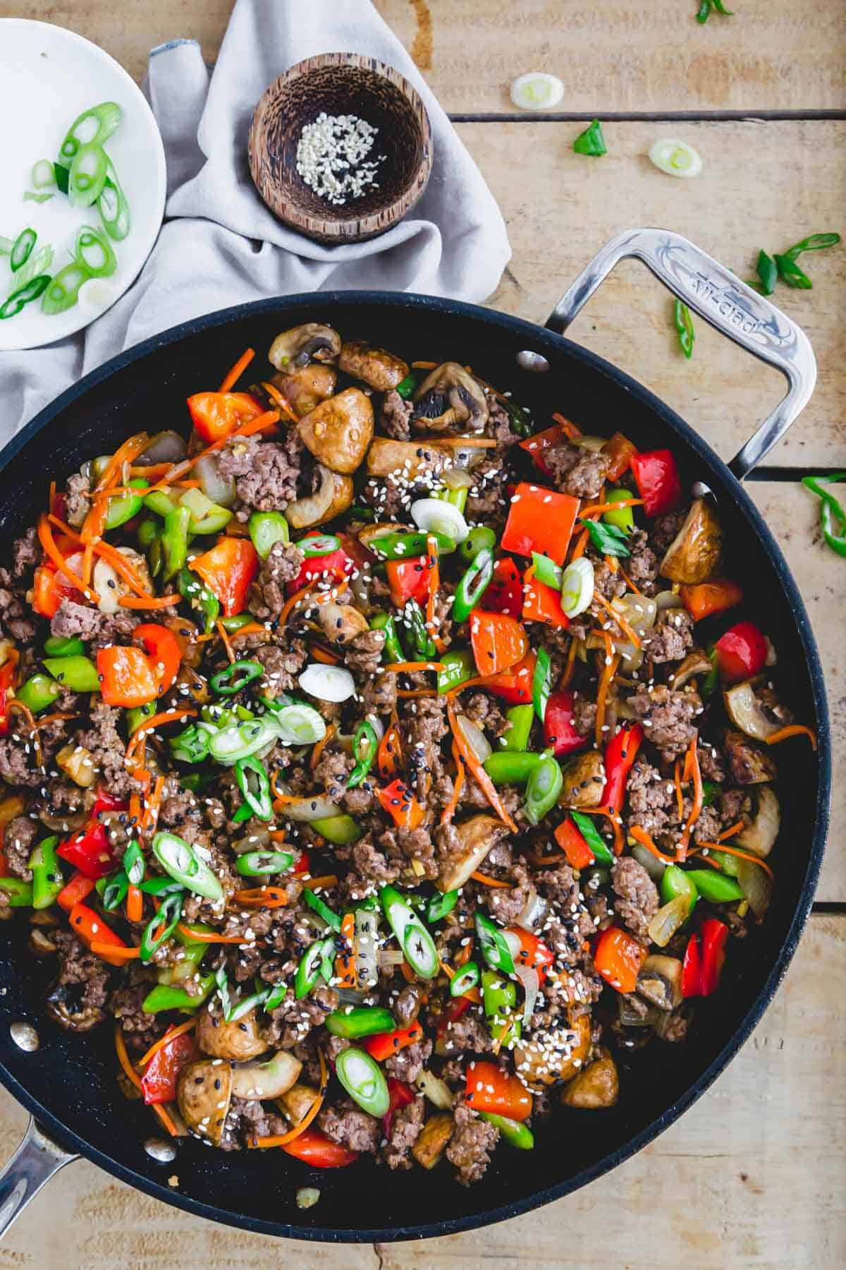 Ground beef stir fry with fresh vegetables in a black pan with sesame seeds and green onion garnish.