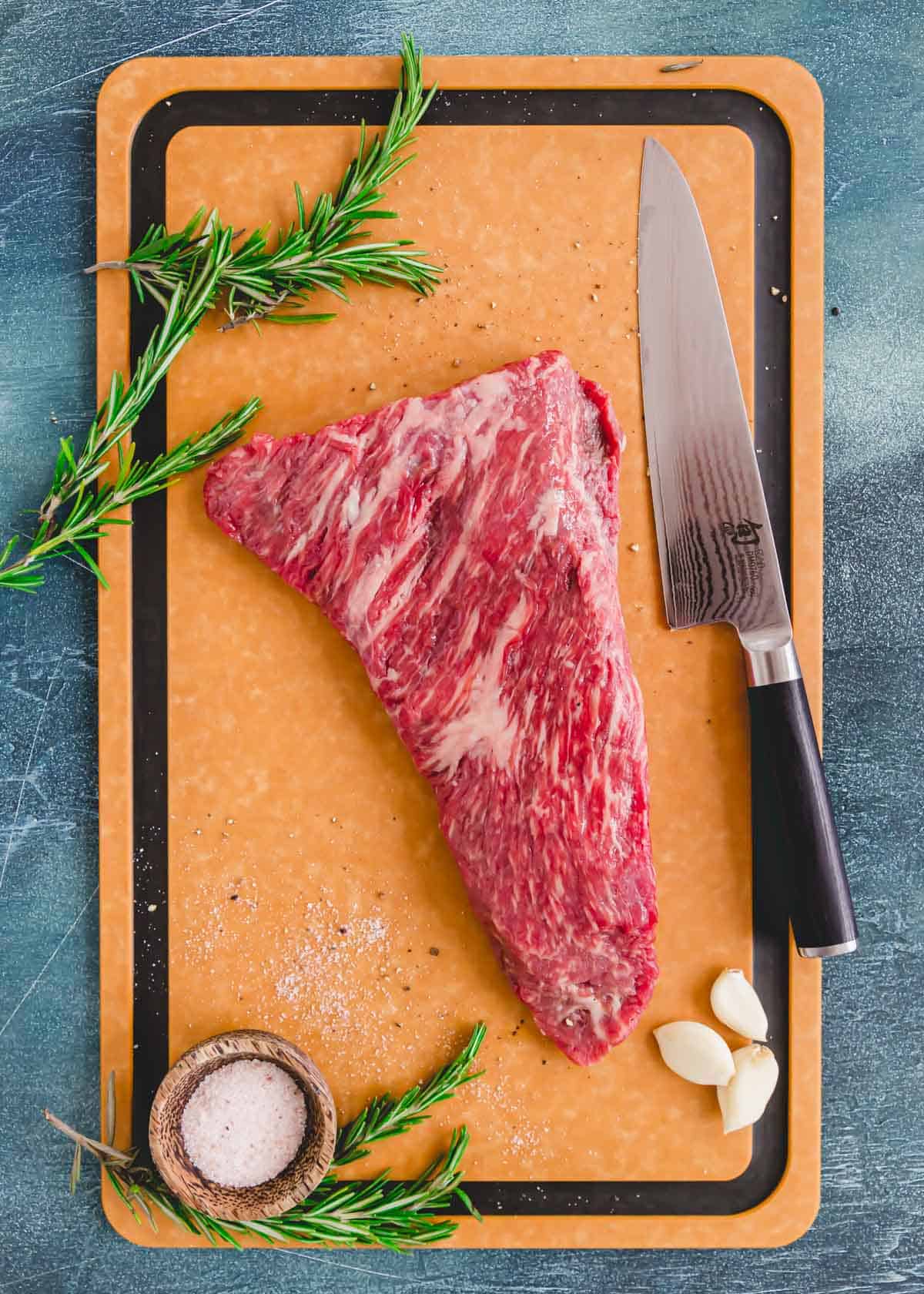 Raw tri tip steak on a cutting board with rosemary, garlic and a chef's knife.