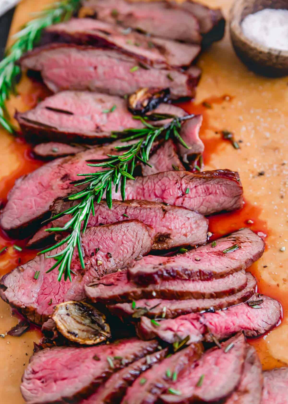 Perfect reverse seared tri tip thinly sliced on a cutting board with rosemary sprigs.