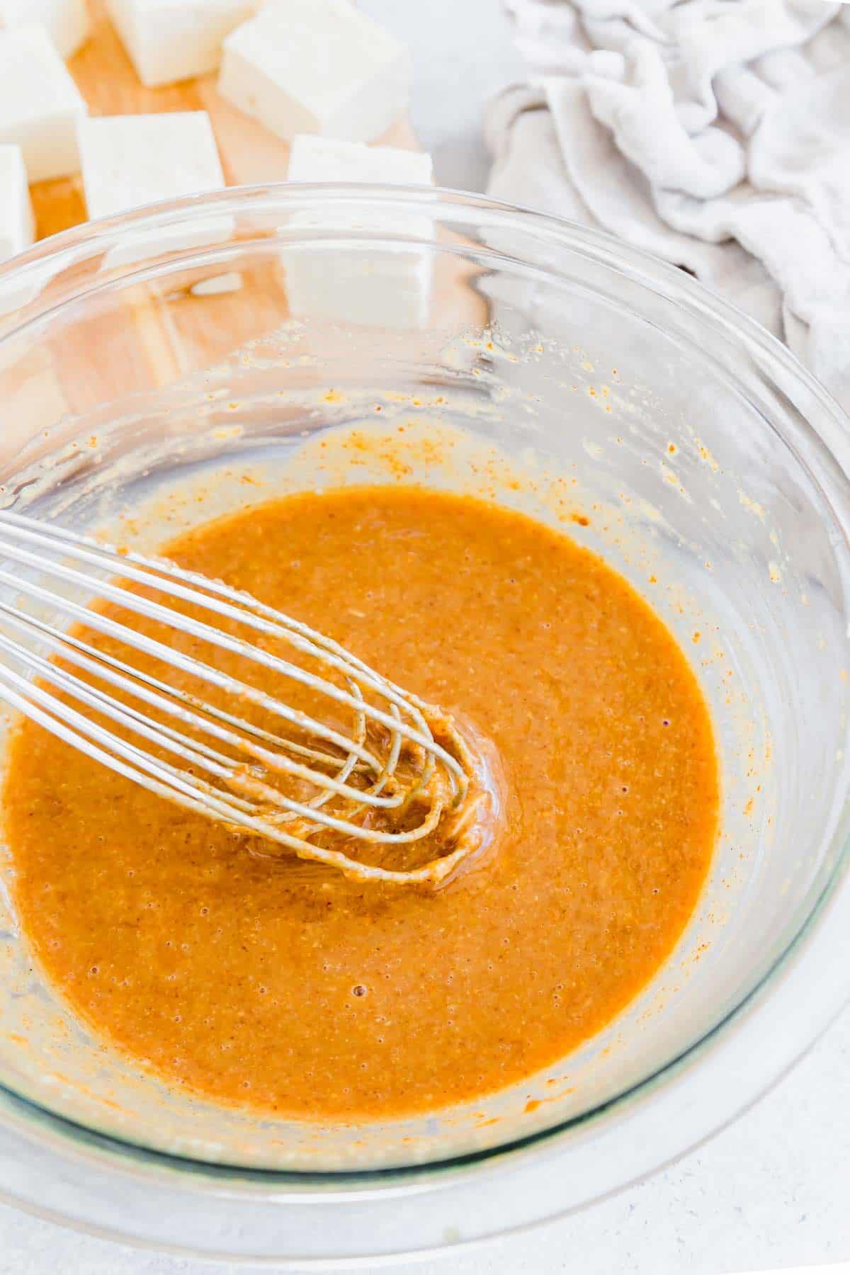 Whisking creamy almond butter tofu marinade in a glass bowl.