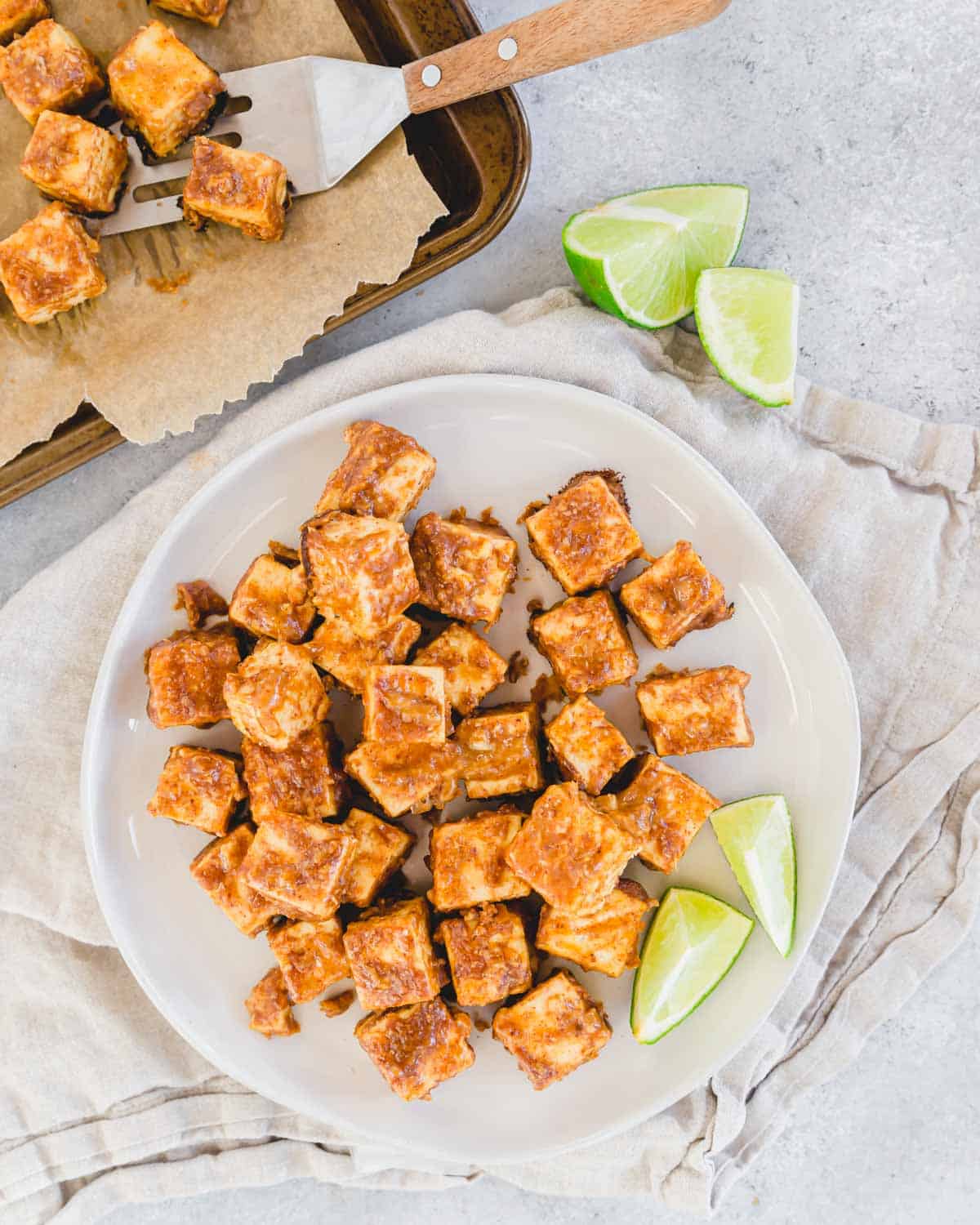 Crispy marinated tofu on a plate with lime wedges. Baking sheet with more tofu and spatula in the background.