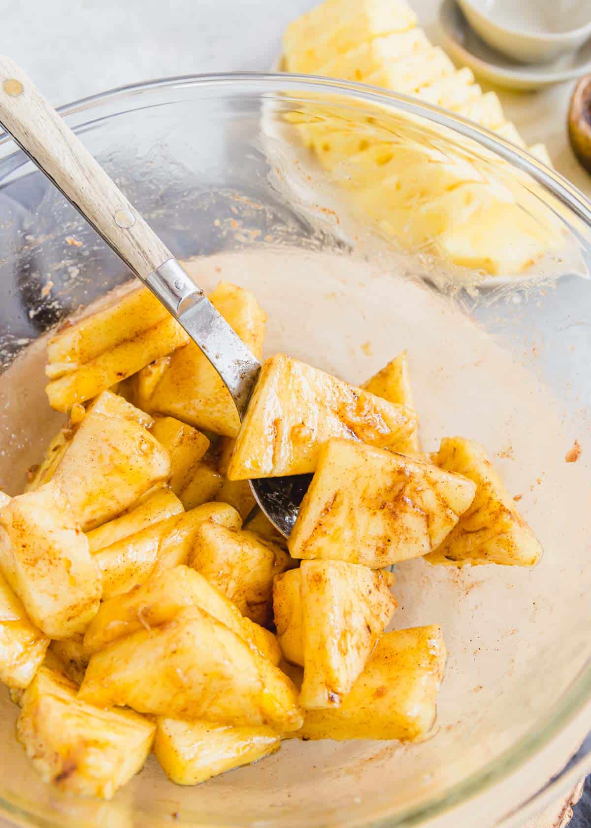 Tossing chopped pineapple with coconut oil, maple syrup and spices in a glass bowl.