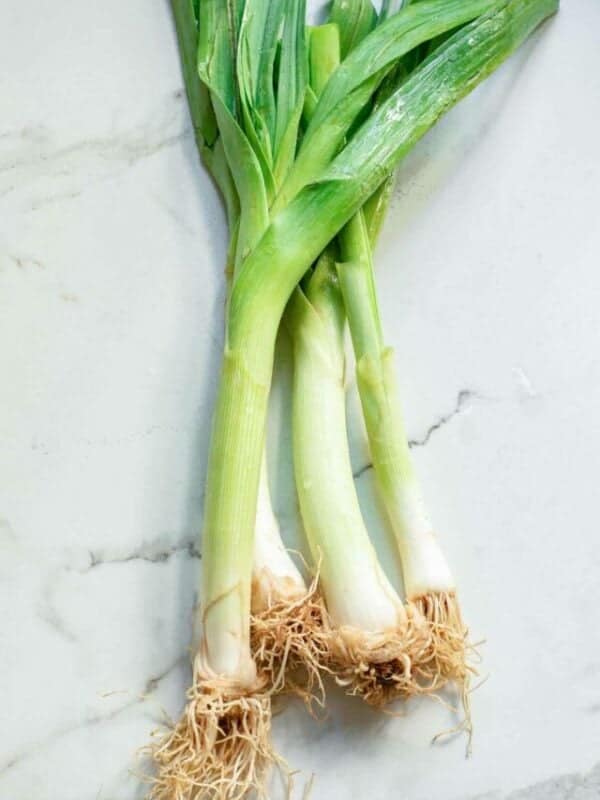 Three fresh leeks with roots intact placed on a marble surface.