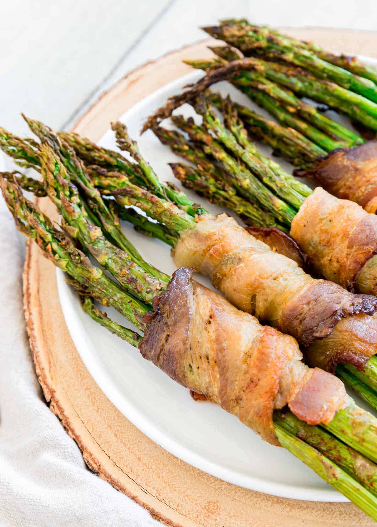Air fryer bacon wrapped asparagus bundles on a white plate with wooden surface beneath.