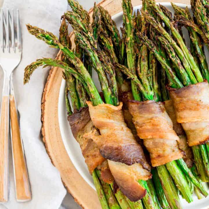 Air fried asparagus bunches wrapped in crispy bacon on a plate with forks to the side.
