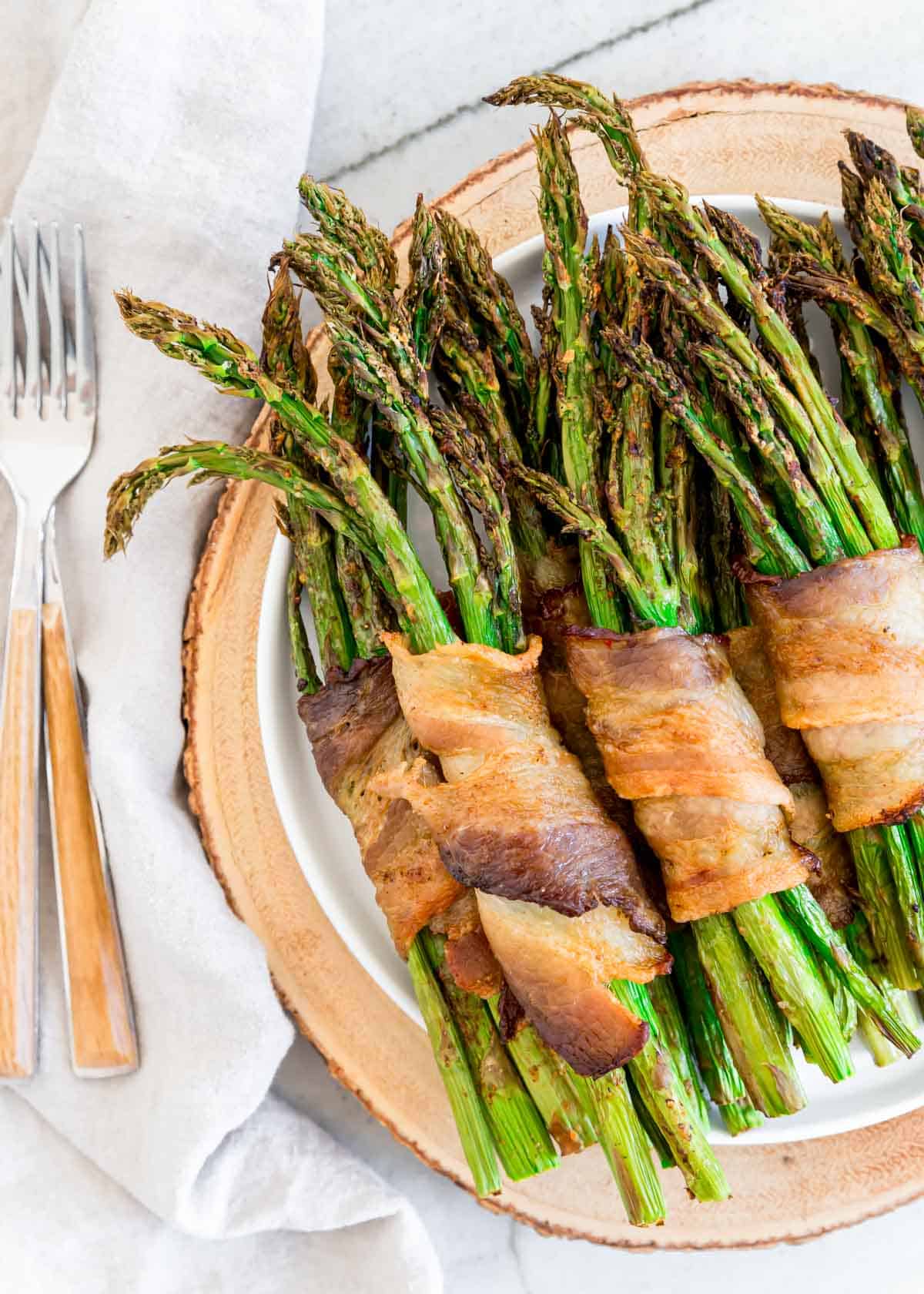 Air fried asparagus bunches wrapped in crispy bacon on a plate with forks to the side.