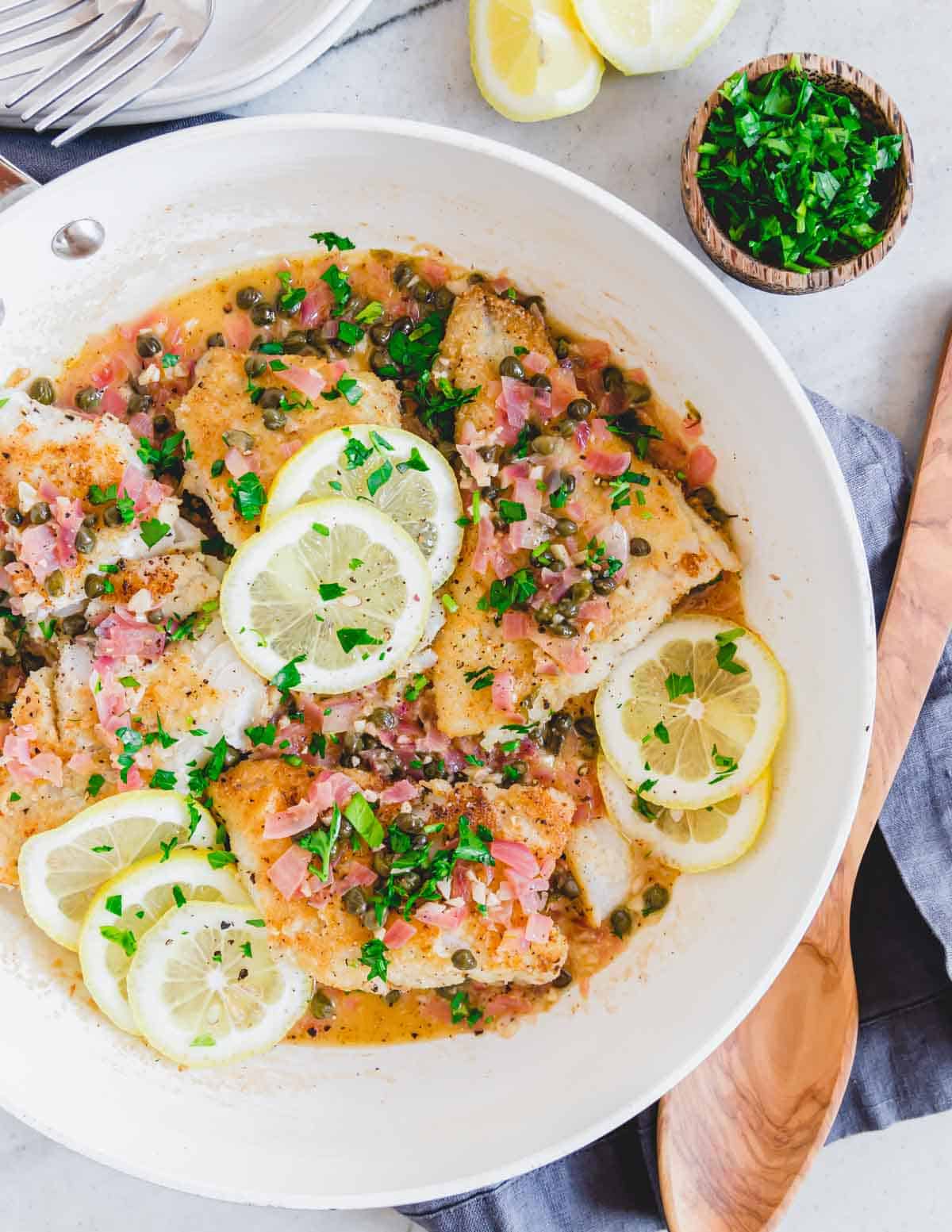 Overhead shot of cod piccata in a skillet with lemon slices and parsley garnish.