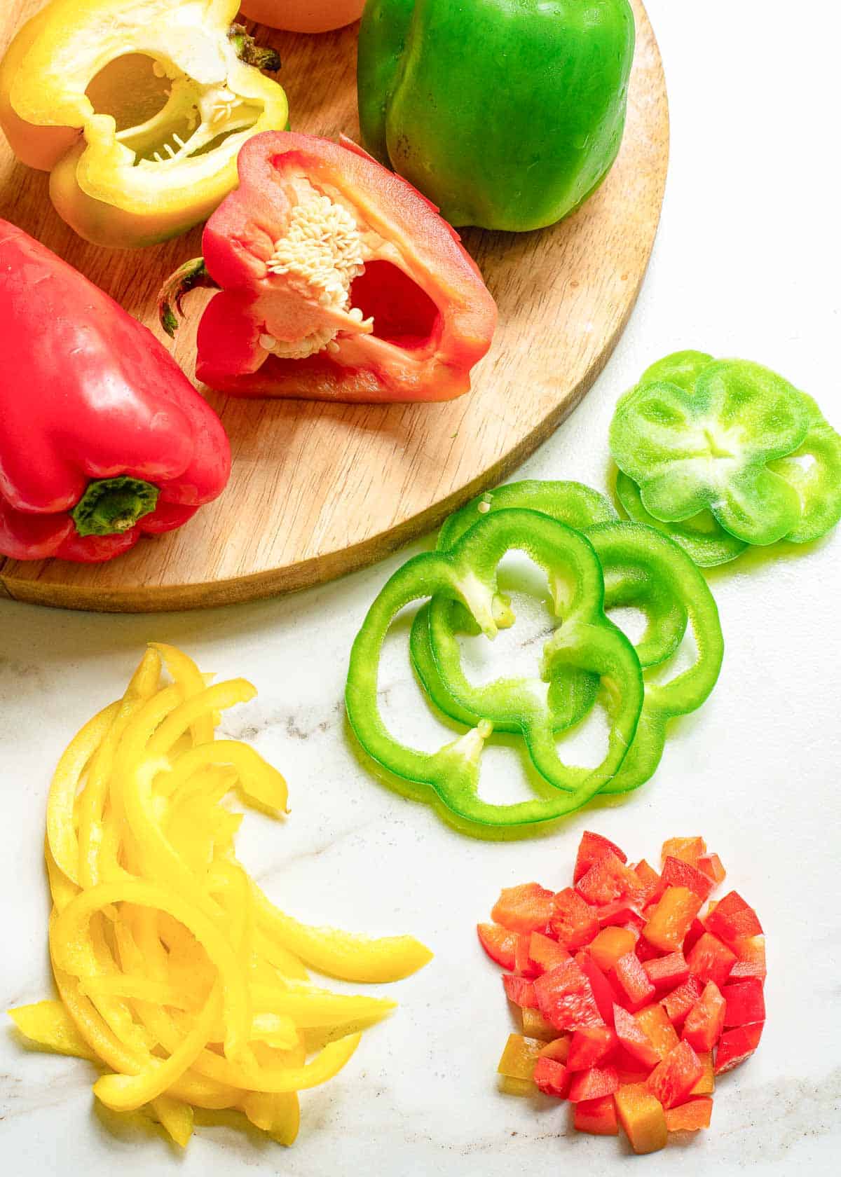 Different color bell peppers cut in a variety of ways on a white marble surface.