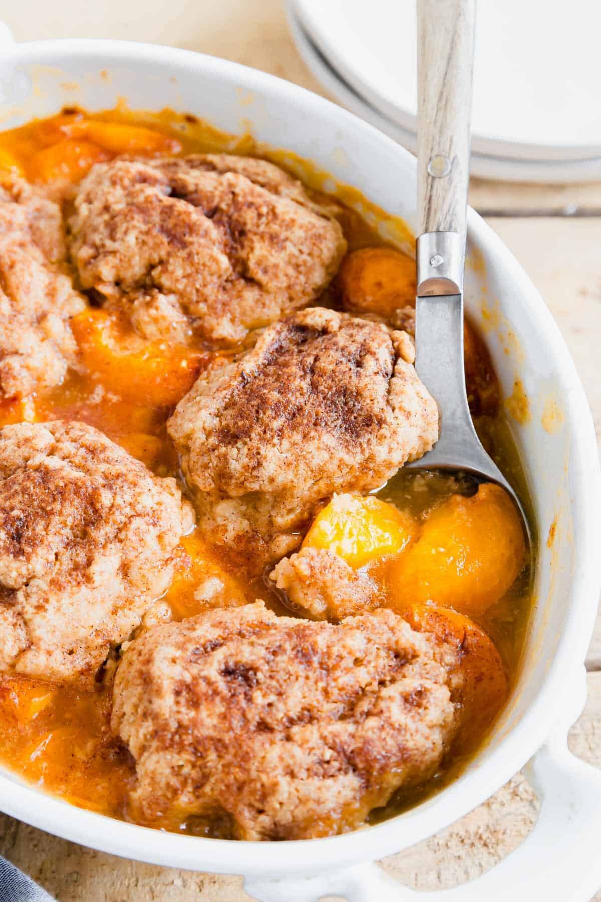 Baked apricot cobbler with wooden and metal serving spoon in a white baking dish.