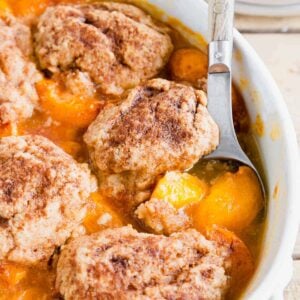 Baked apricot cobbler with wooden and metal serving spoon in a white baking dish.