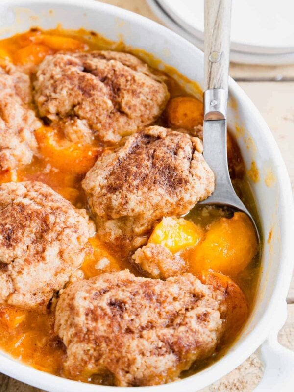 Baked apricot cobbler with wooden and metal serving spoon in a white baking dish.