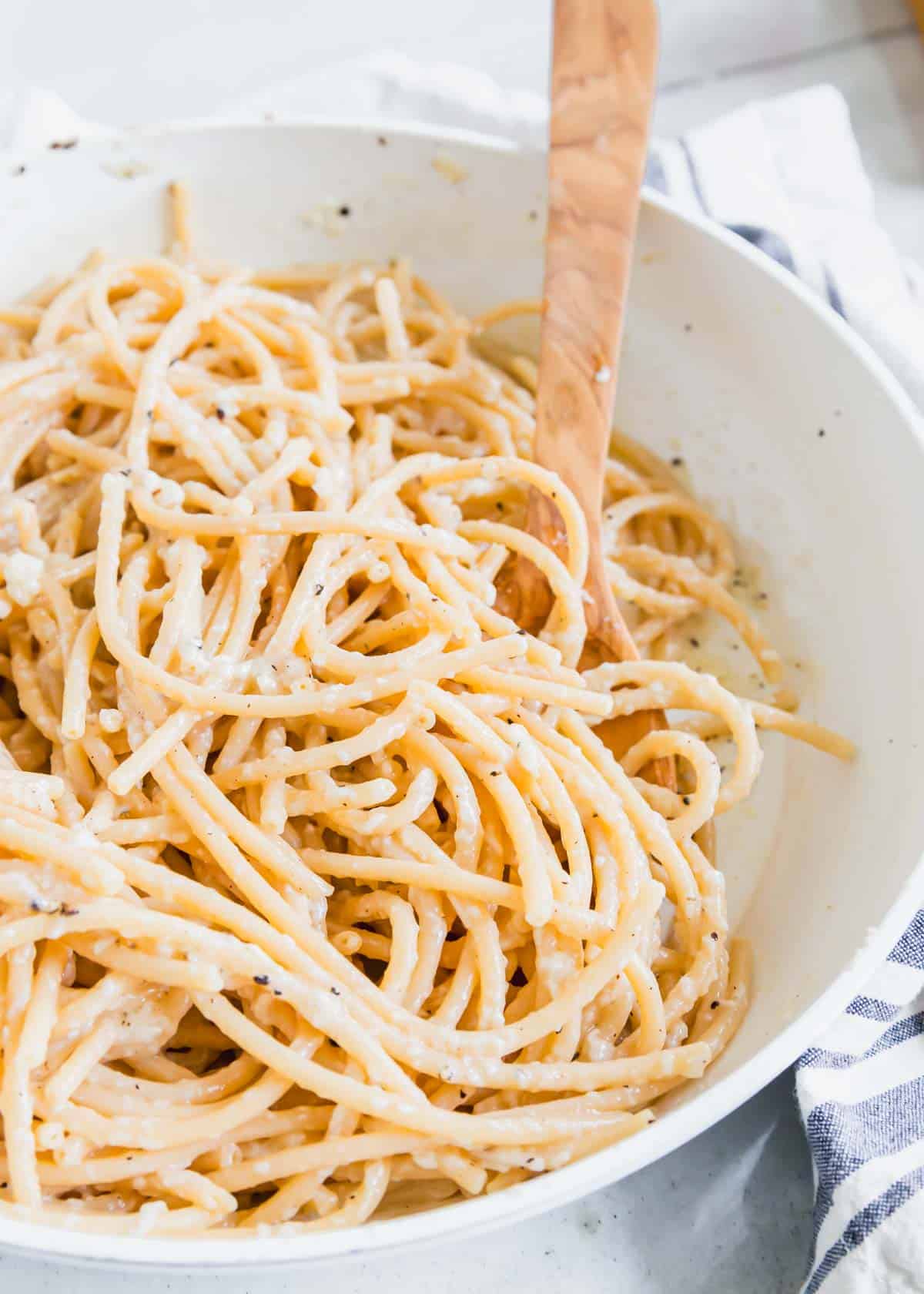 Bucatini cacio e pepe in a white skillet with a wooden serving spoon.