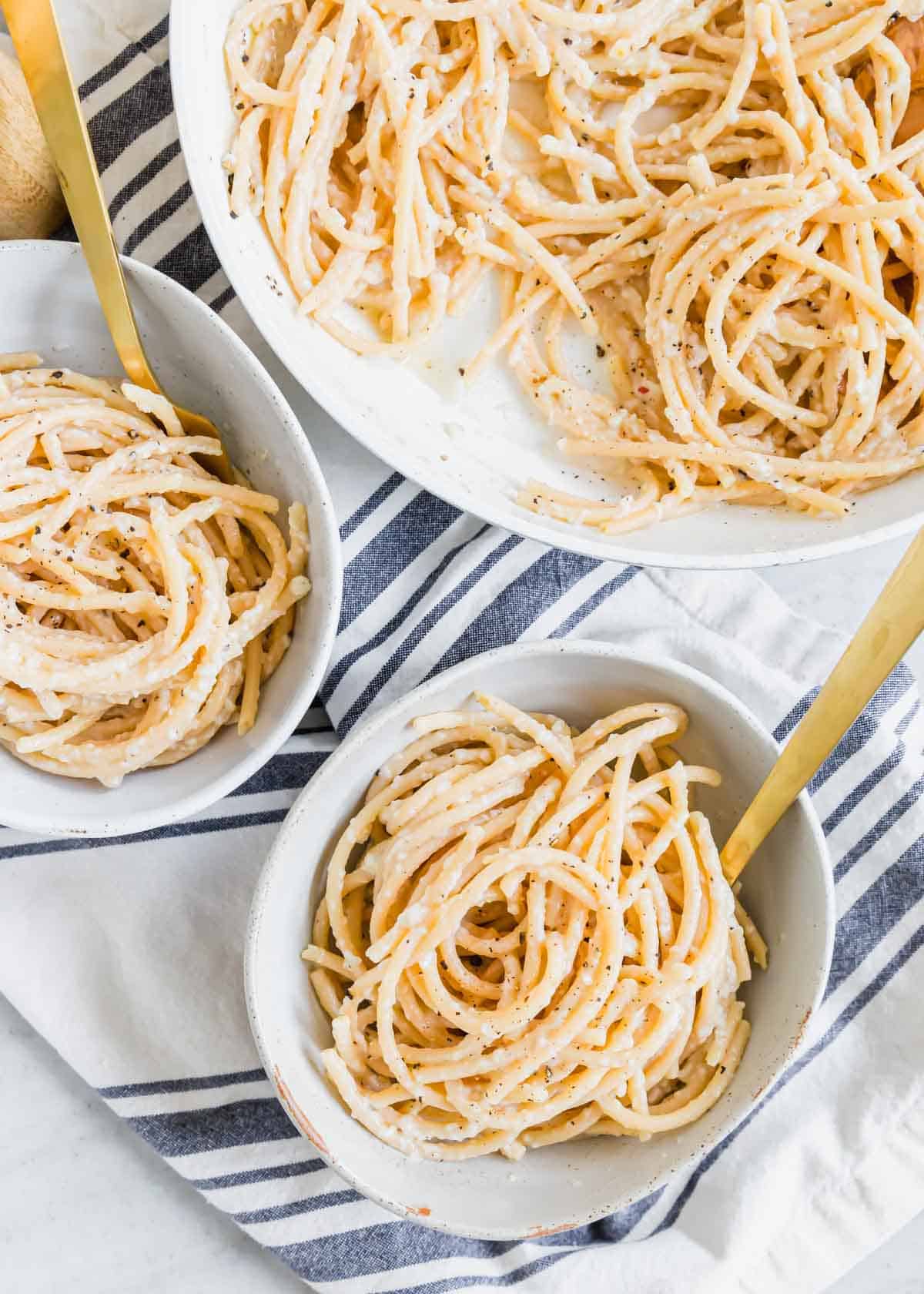 Bucatini cacio e pepe in bowls with gold forks and a skillet in the background.