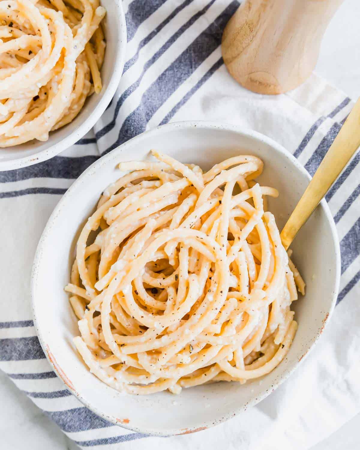 Overhead of bucatini pasta cacio e pepe in a pottery bowl with a fork and a pepper mill in the background.